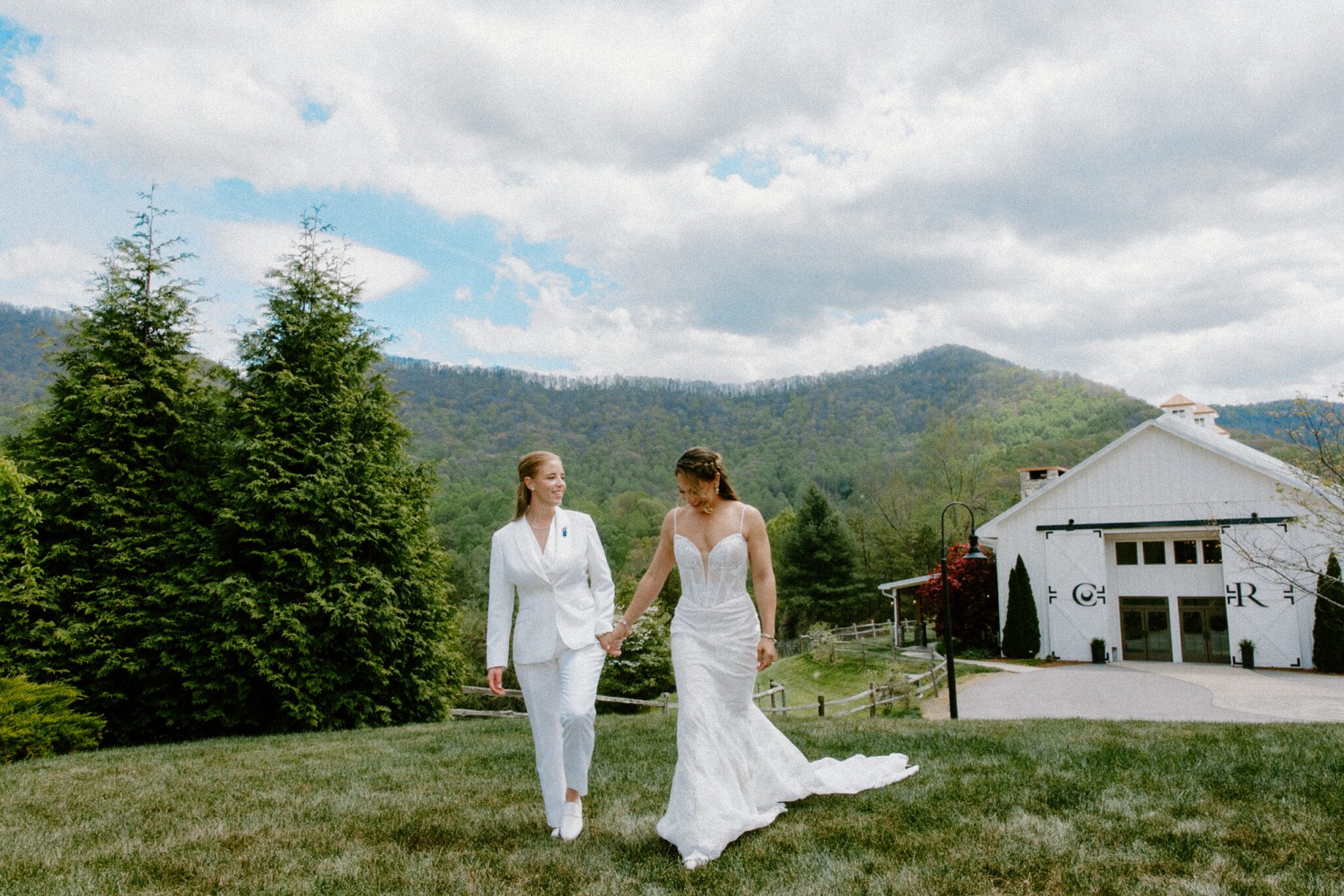 Two brides, one in a white suit and the other in a white wedding dress, hold hands while walking on grass with a barn and tree-covered mountains in the background.