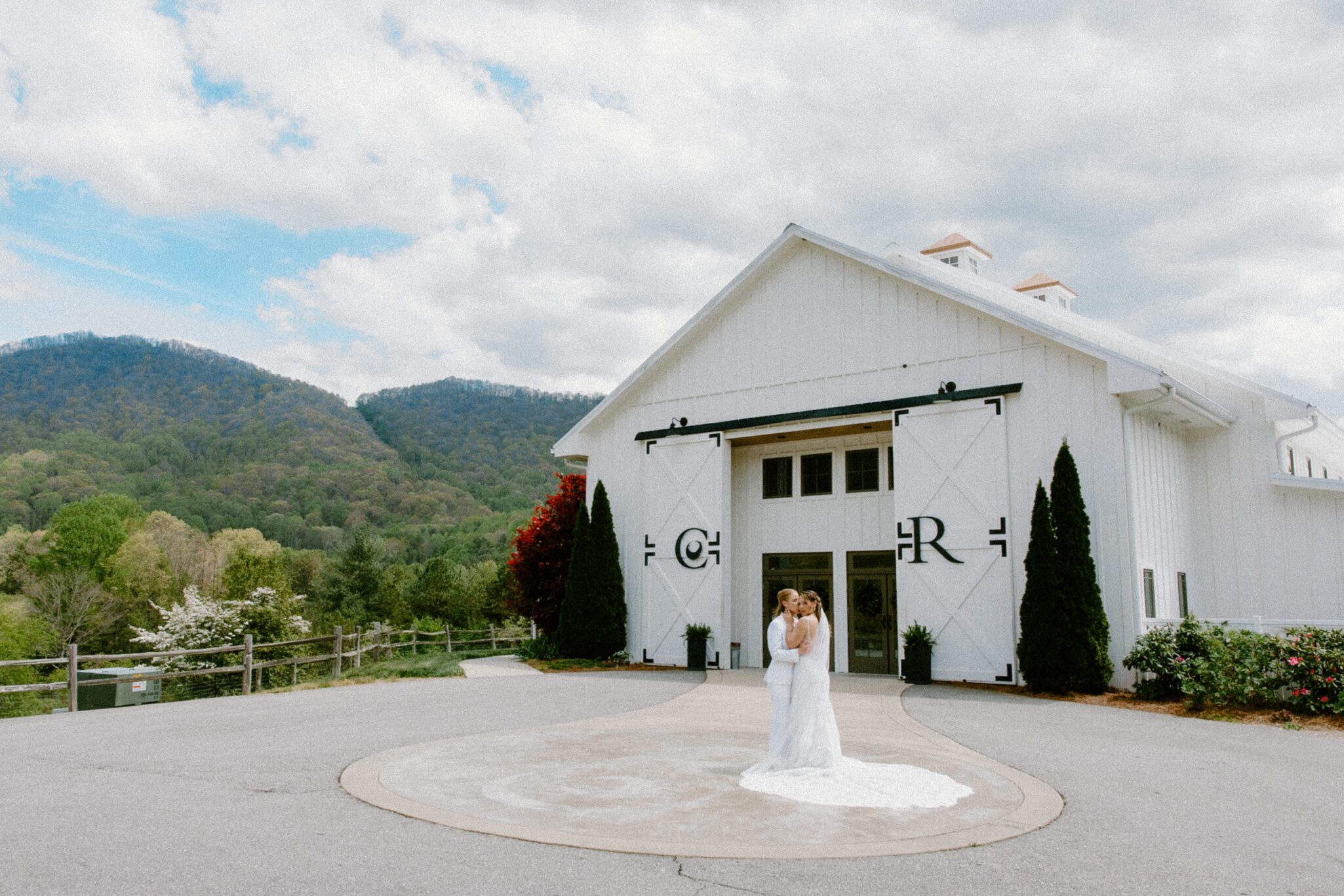Two brides in white gowns embrace and smile outside a large, modern white barn with black doors, surrounded by trees and mountains under a partly cloudy sky.