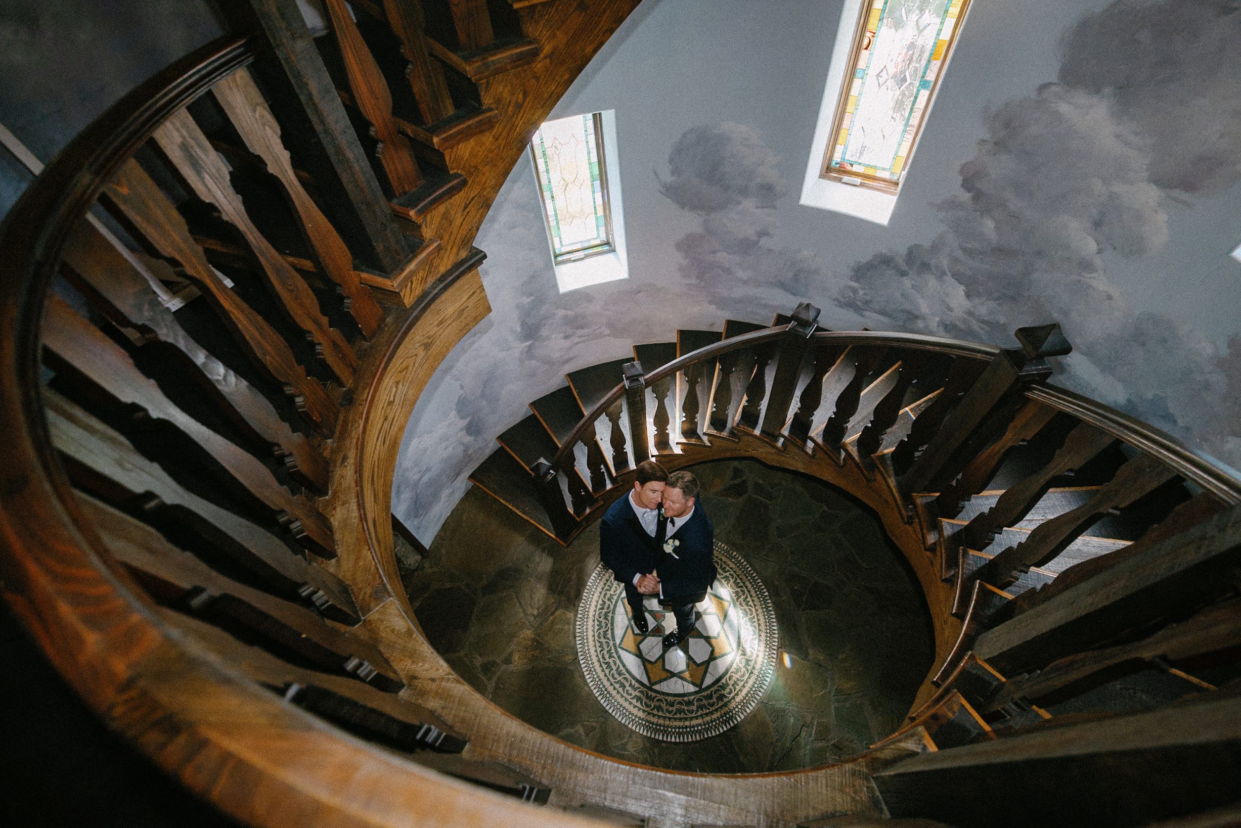 A couple dressed formally stands at the center of a circular patterned floor, embracing, at the bottom of a spiral wooden staircase surrounded by stained glass windows during their enchanting Castle Ladyhawke wedding.