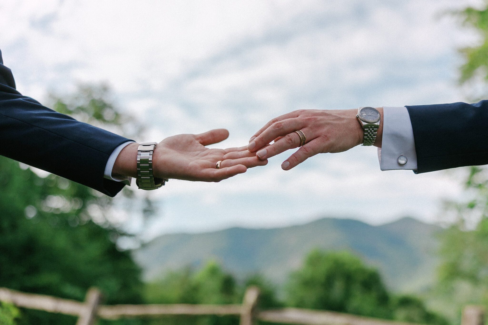 Two people in suits reach toward each other with extended hands, one wearing a wedding ring, against a blurred outdoor background with trees and mountains—evoking the romantic atmosphere of a Castle Ladyhawke wedding.