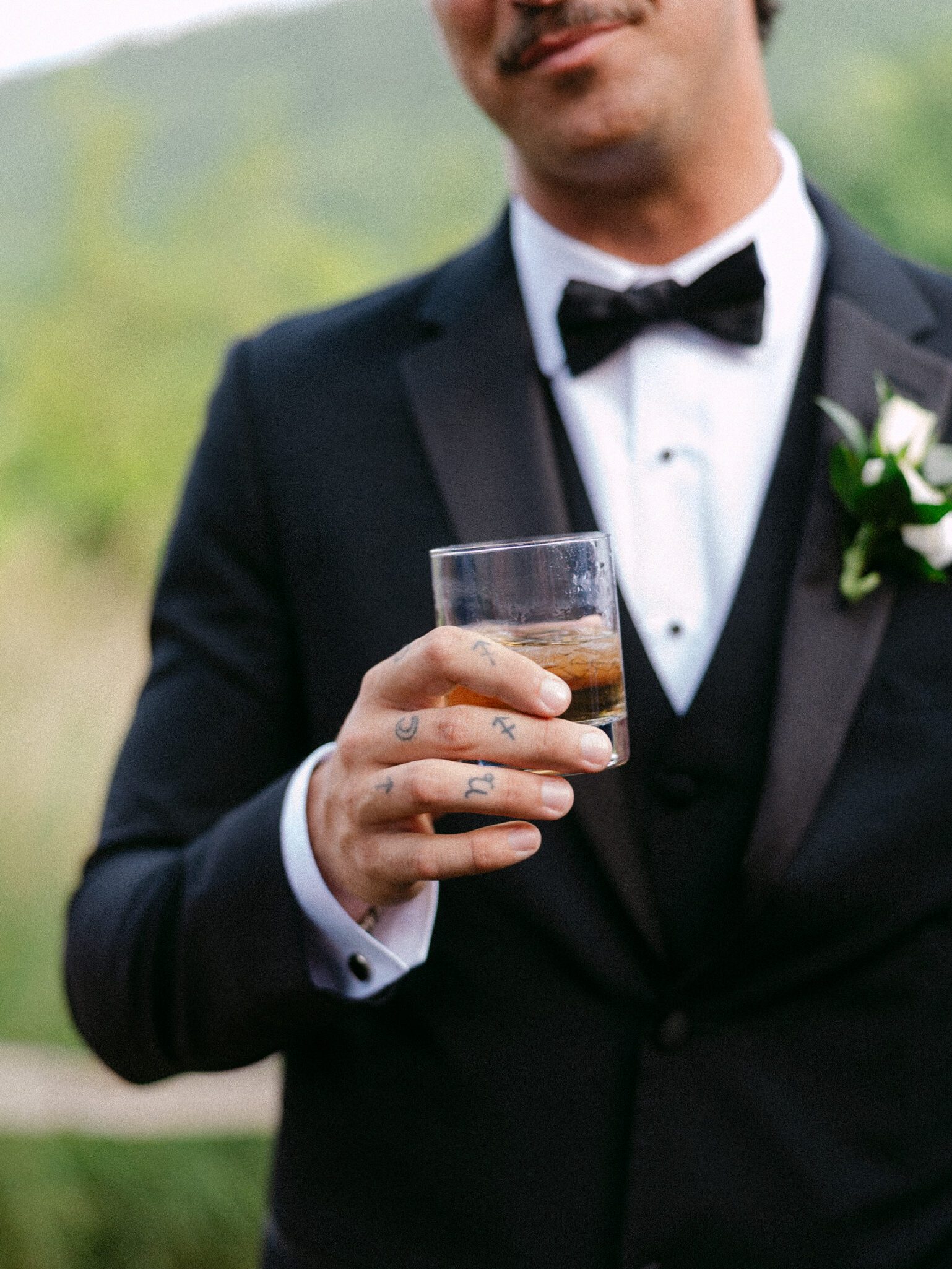 A man in a black tuxedo holds a glass of whiskey. He has tattoos on his fingers and a boutonniere on his lapel. The background is blurred greenery.