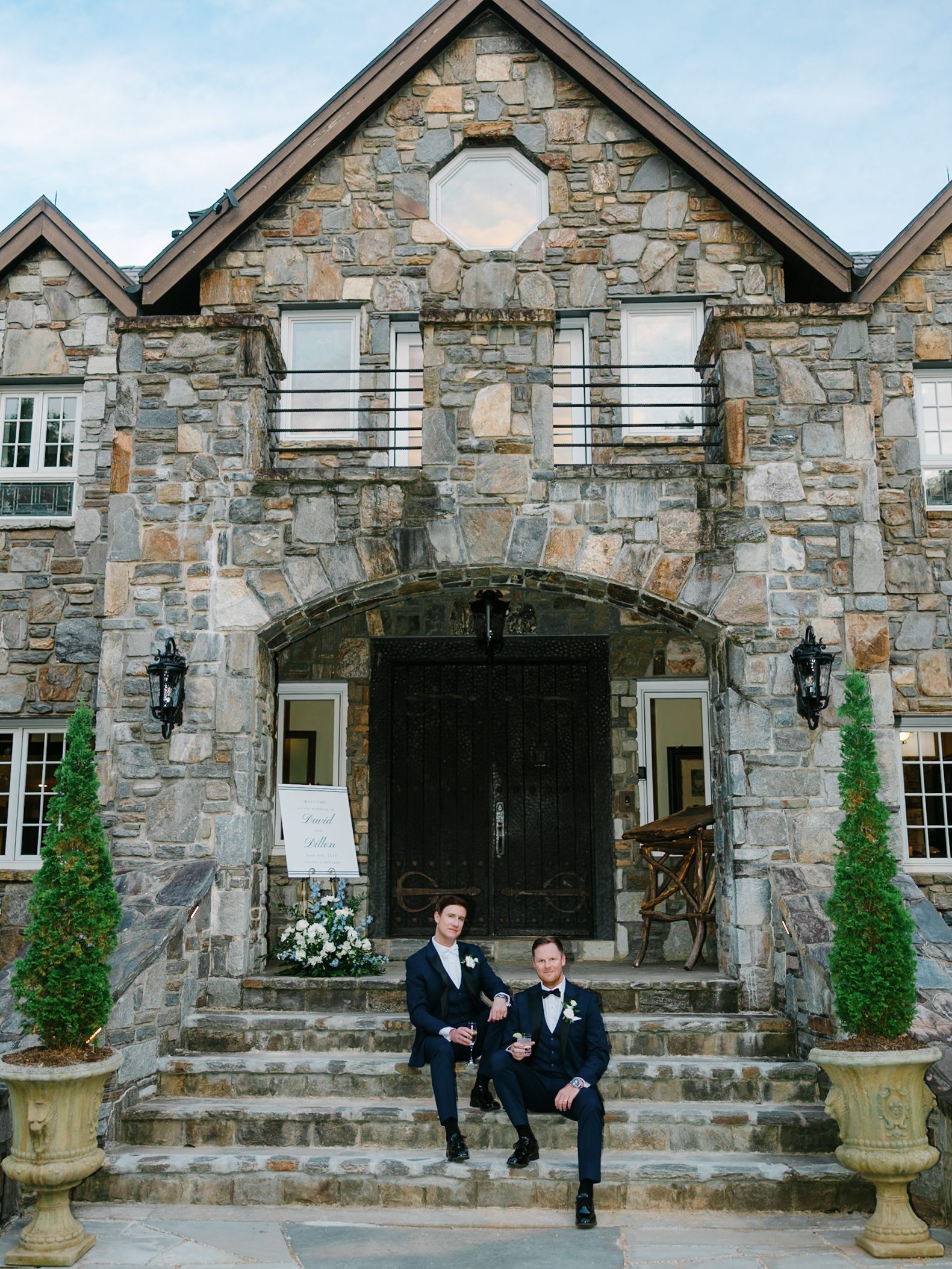 Two men in suits sit on stone steps in front of a large stone building with double doors and arched entryway, flanked by two tall potted plants. A floral arrangement and welcome sign are near the doors.