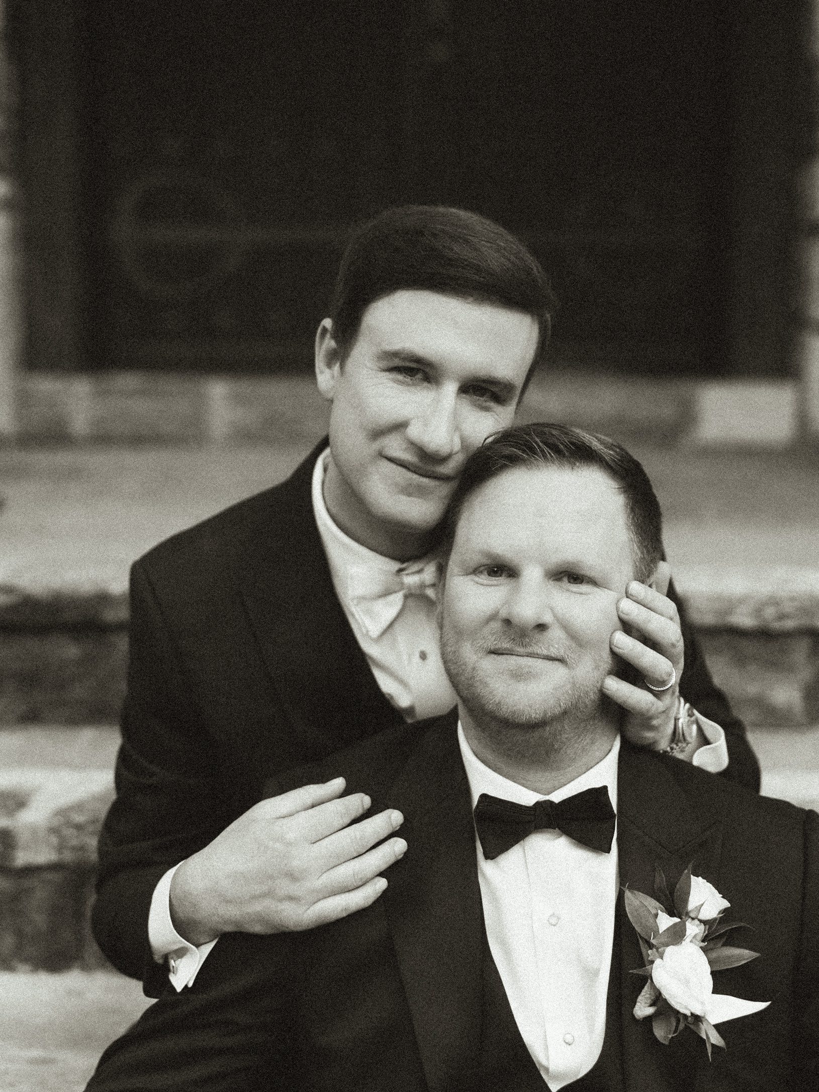 Two men in tuxedos sit closely together on stone steps, smiling softly. One gently rests his hand on the others cheek. Both wear bow ties and boutonnieres, suggesting a wedding or formal occasion. The image is in black and white.