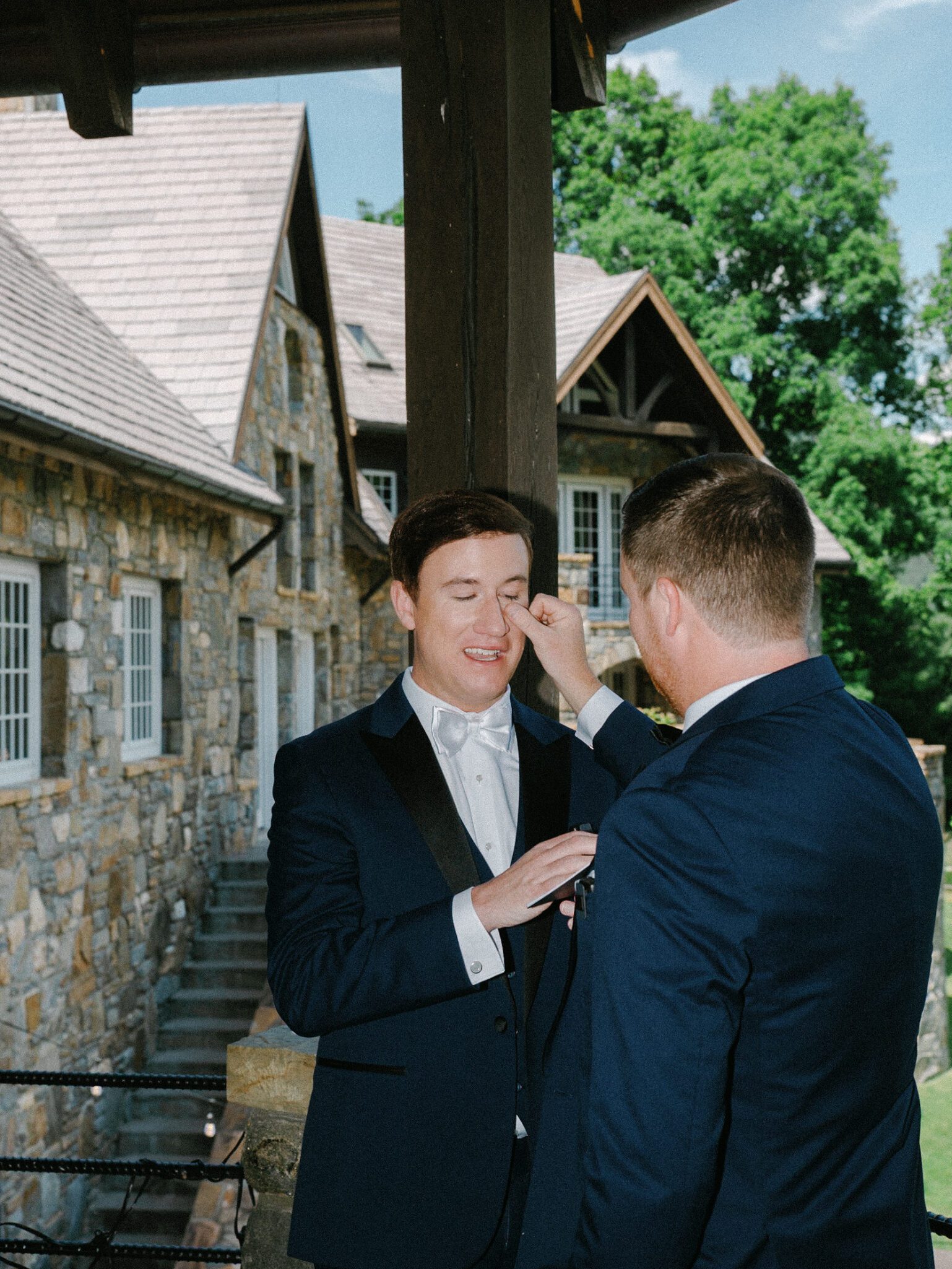 Two men in formal suits stand outside the stone walls of Castle Ladyhawke on their wedding day. One gently wipes the other's face, possibly fixing his makeup, while both smile. Trees and a bright sky are visible in the background.