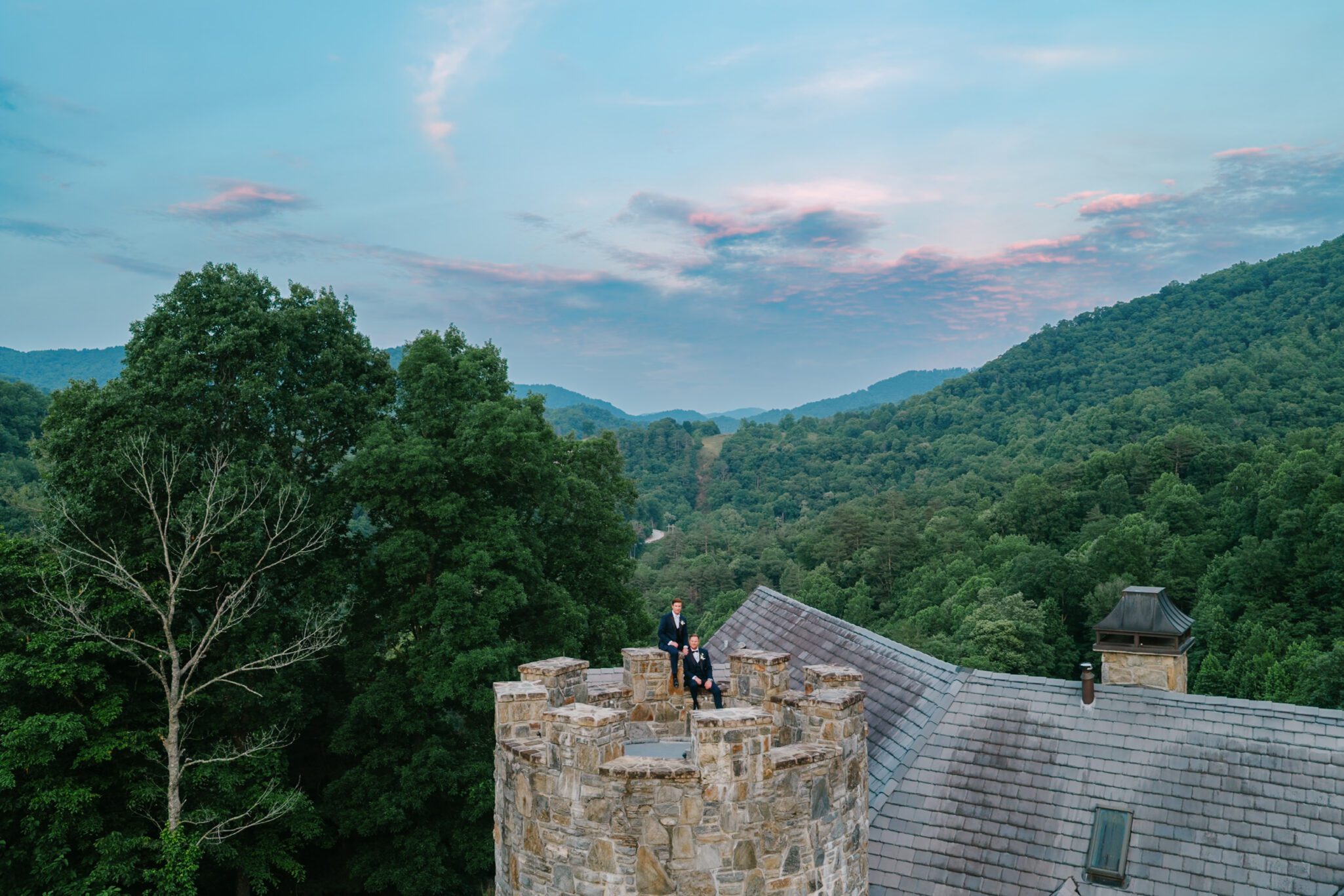 A couple in formal attire stands atop a stone castle turret during a drone shot at a Castle Ladyhawke wedding, surrounded by green forested hills at sunset, with pink clouds and mountains in the distance.