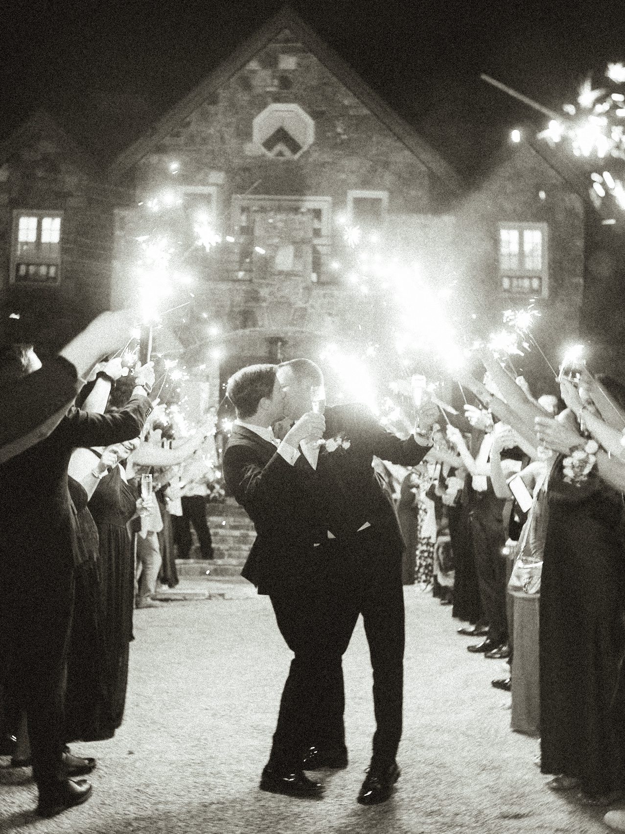 Two people in suits kiss passionately while guests form a tunnel around them, holding sparklers aloft. The celebration takes place outdoors at night in front of the grand stone facade of a Castle Ladyhawke wedding.