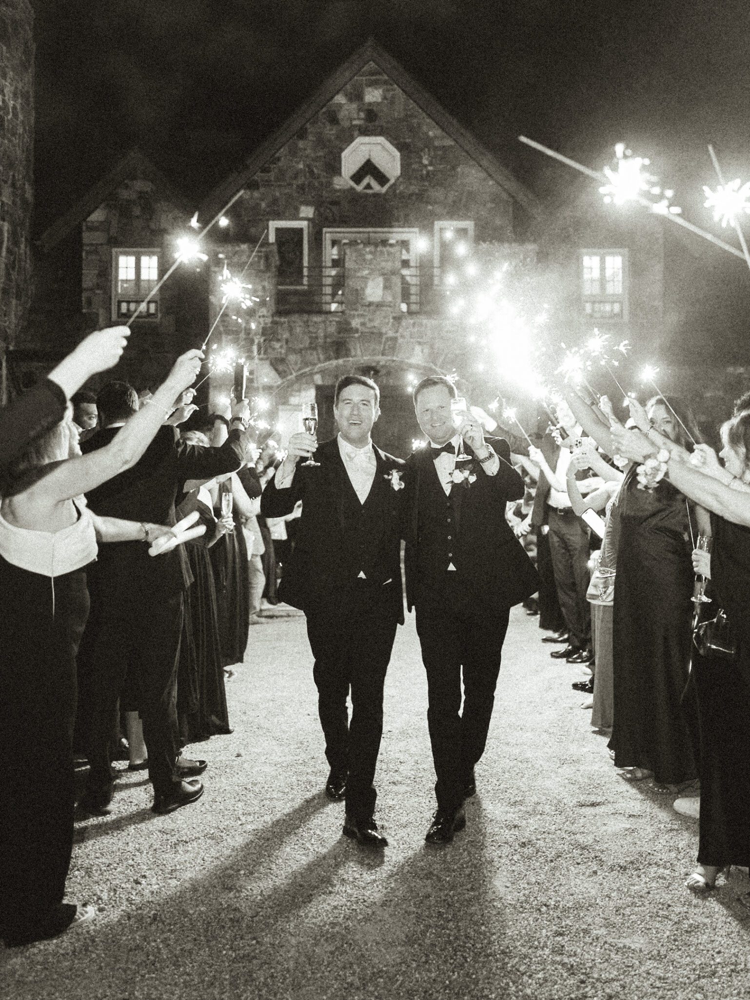 Two grooms in suits walk hand-in-hand, smiling, surrounded by guests holding sparklers at night in front of a stone building. The scene is festive and celebratory.