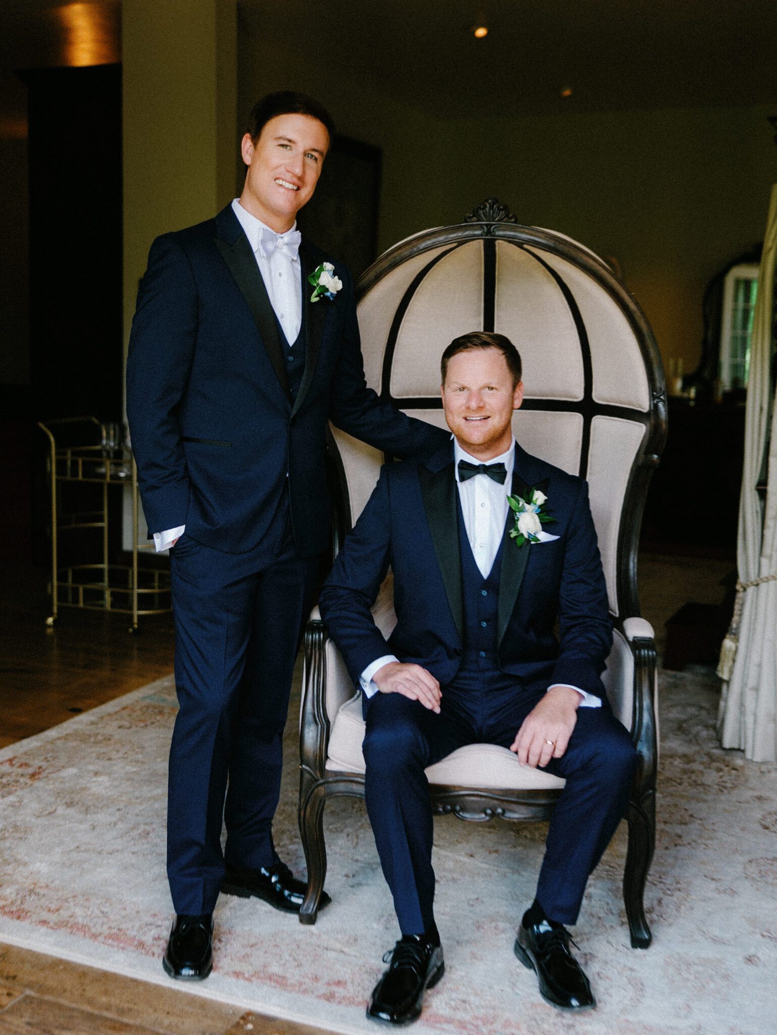 Two men in dark suits and bow ties pose indoors; one is seated on an elegant chair, while the other stands with a hand on the chair back. Both wear boutonnieres and smile at the camera.