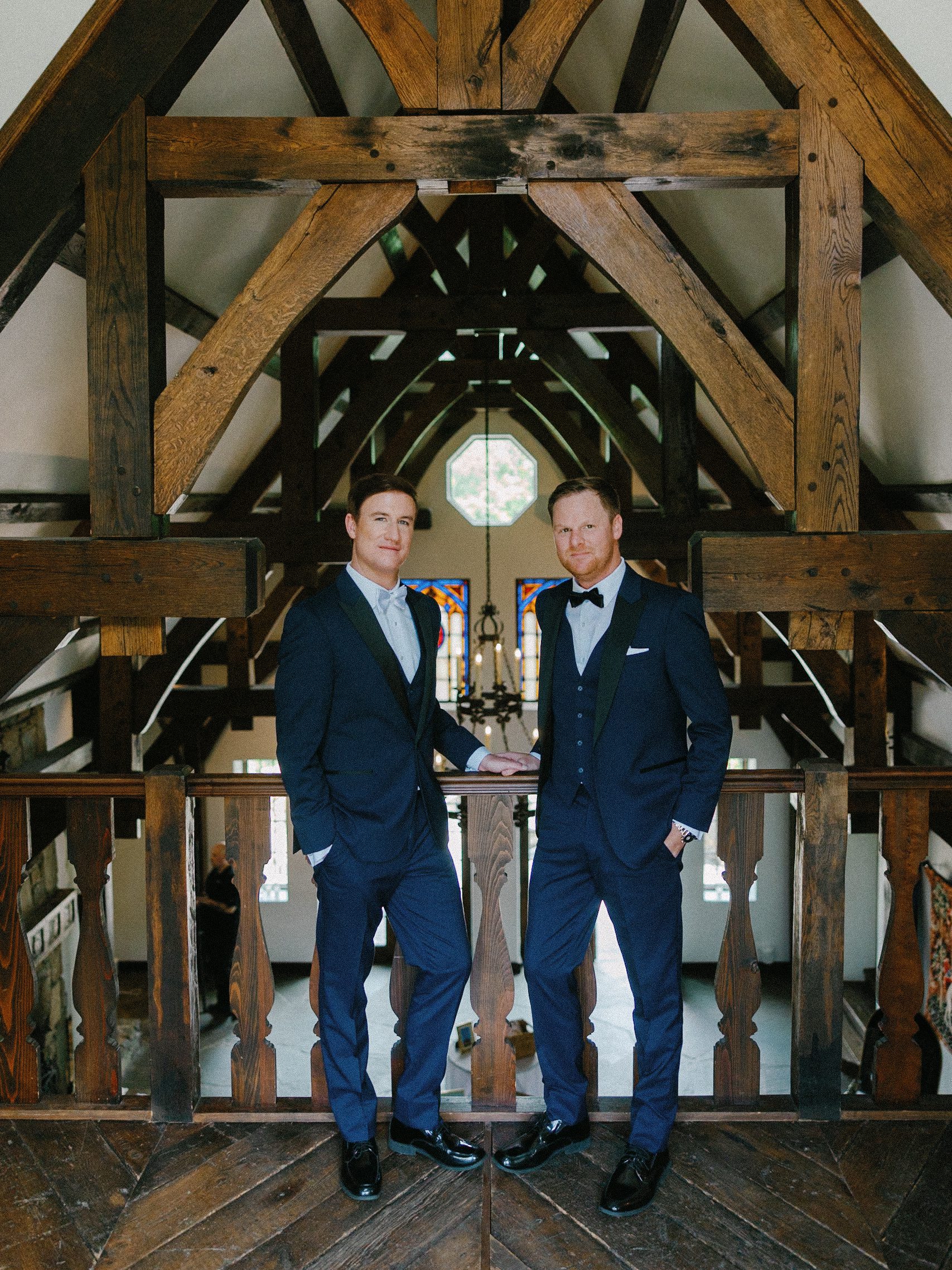 Two men in suits stand confidently on a wooden balcony inside a building with exposed beams, stained glass windows, and natural light streaming in from above.