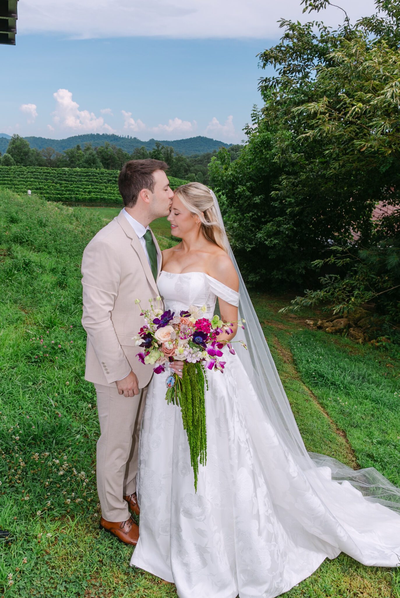 A groom in a tan suit kisses the brides forehead as she holds a bouquet of purple, cream, and pink flowers. They stand outdoors on green grass with trees and mountains in the background.