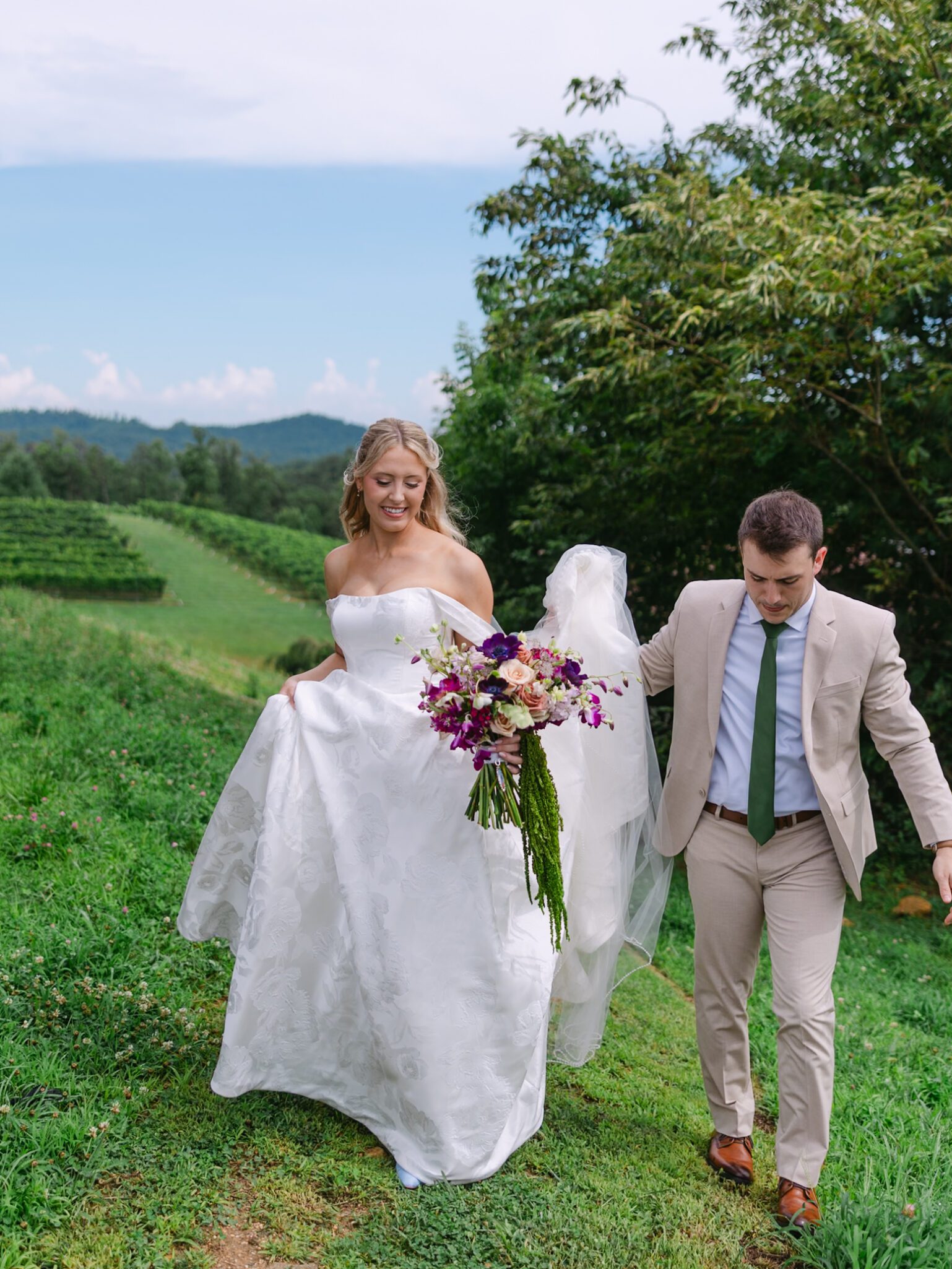 A bride in a white dress holds a bouquet and lifts her gown while walking outdoors beside a man in a beige suit, both surrounded by green grass and trees with hills in the background.