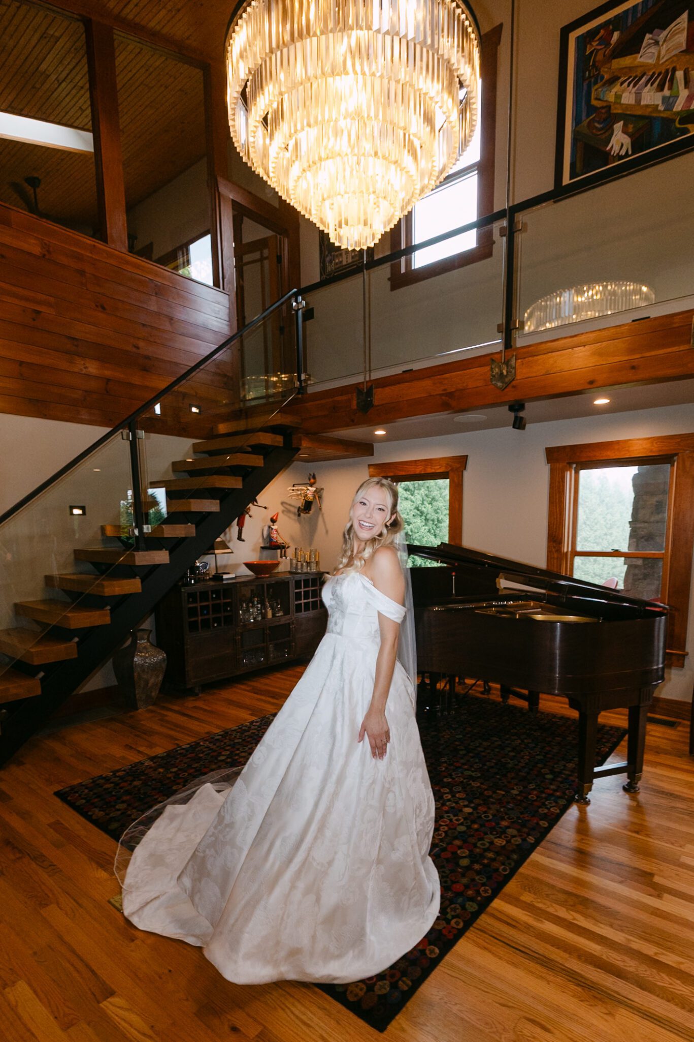 A woman in an off-shoulder white gown stands smiling by a grand piano in a spacious, wood-accented room with a large chandelier and staircase.