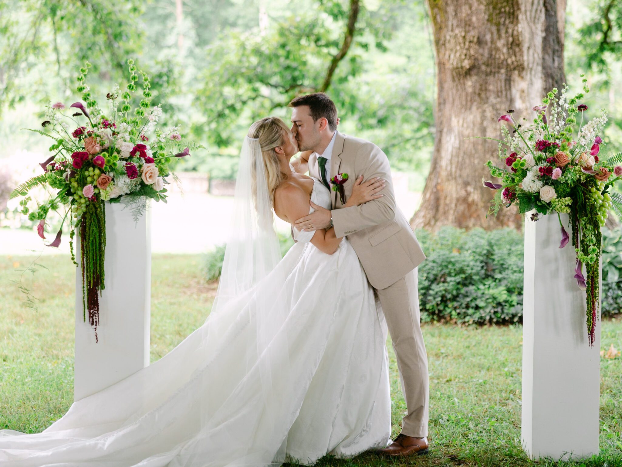 A bride and groom share a kiss outdoors between two large floral arrangements, surrounded by greenery and trees. The bride wears a white gown and veil, and the groom is in a tan suit.