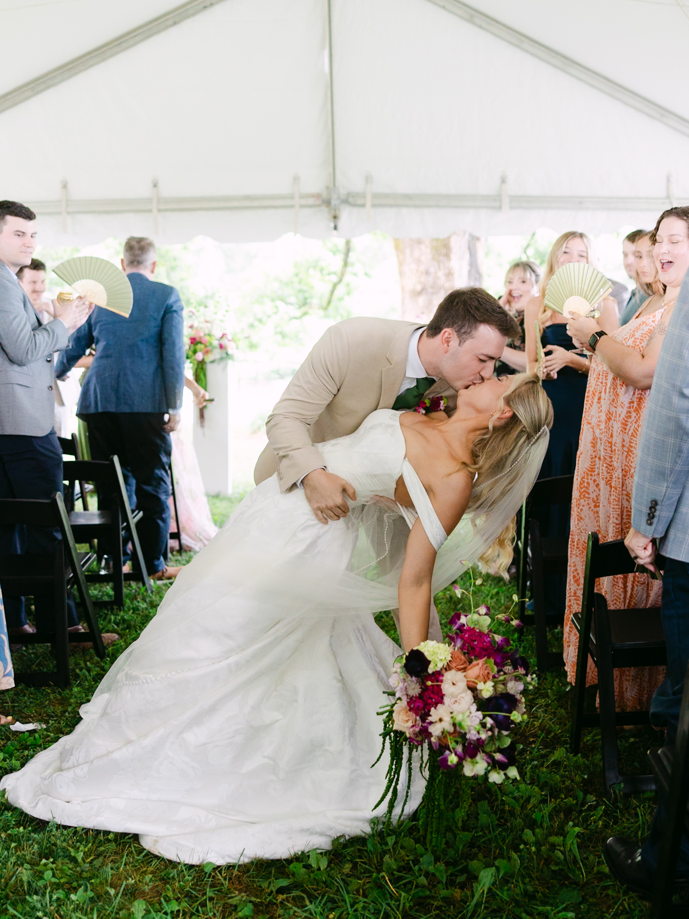A groom dips and kisses his bride at an outdoor wedding ceremony under a tent, surrounded by cheering guests holding handheld fans. The bride holds a vibrant bouquet.