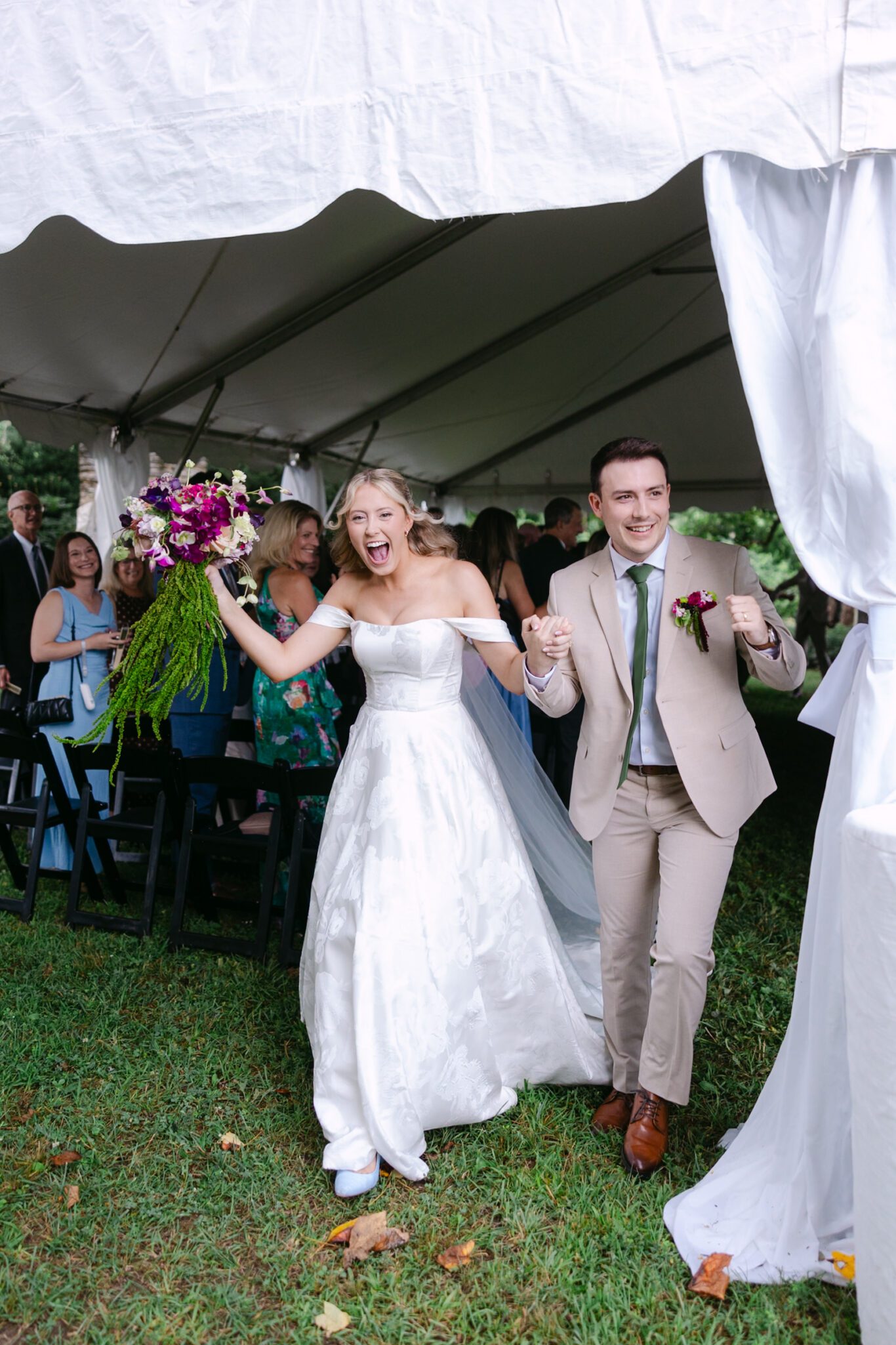 A joyful bride in a white gown and groom in a beige suit walk hand-in-hand under a tent at Marked Tree Vineyards, smiling and celebrating while guests look on. The bride holds a bouquet of purple flowers.