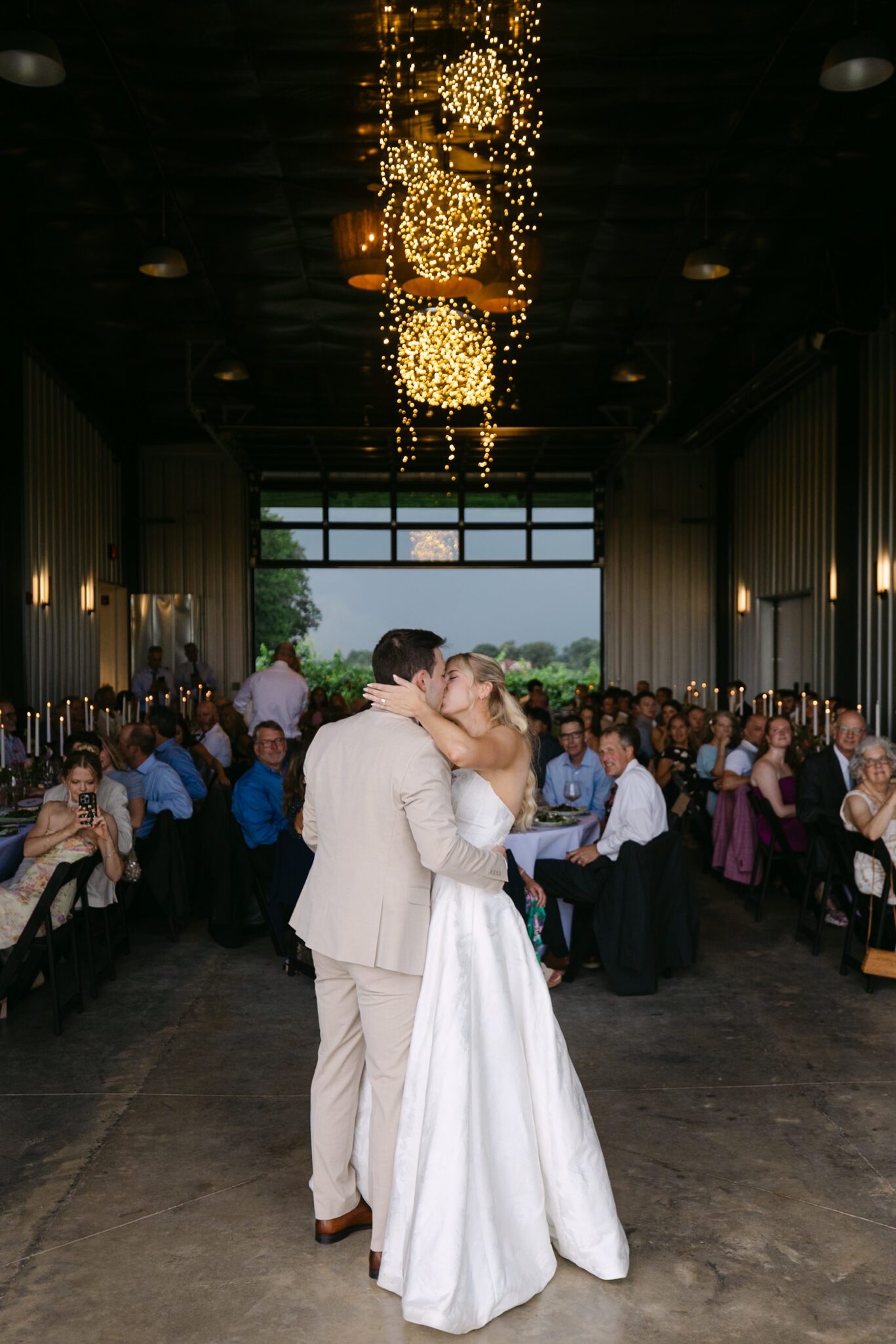 A bride and groom share their first dance in a rustic barn venue, surrounded by seated guests and warm string lights overhead.