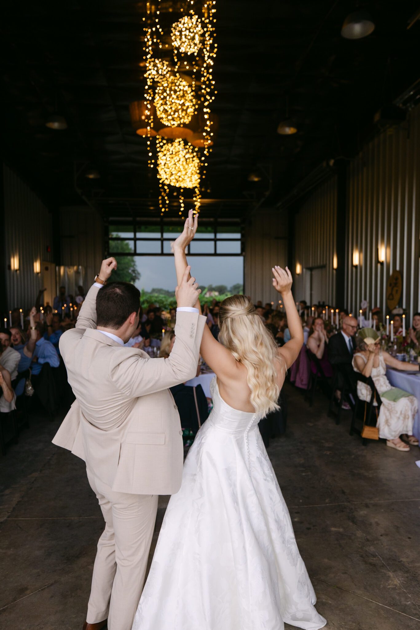 A bride and groom joyfully enter a reception hall, holding hands with arms raised, as guests seated at tables cheer them on. Warm lights hang from the ceiling above.