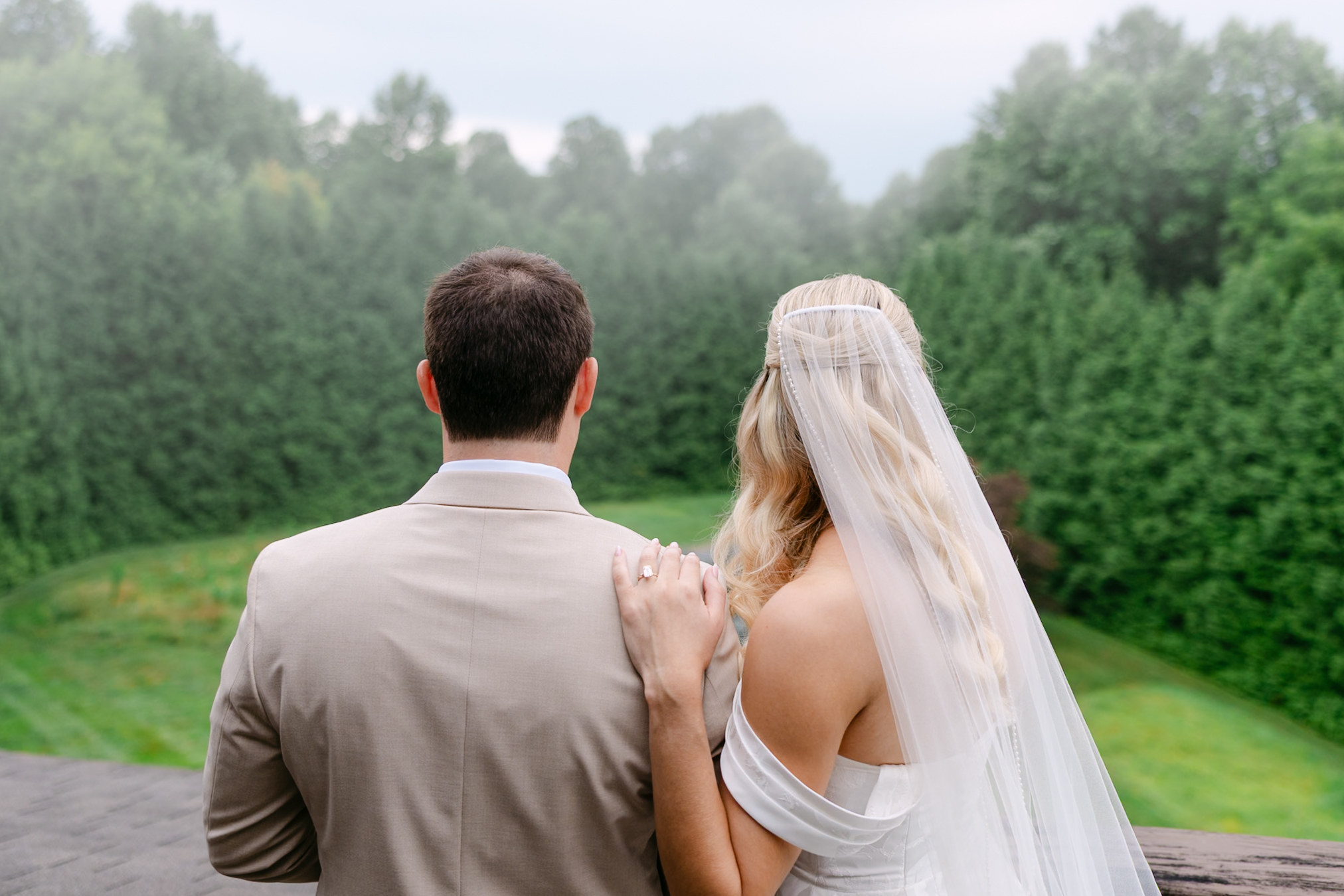 A bride and groom, seen from behind, stand close together outdoors. The bride wears a white dress and veil, and the groom wears a tan suit. Lush green trees and grass fill the scenic background.