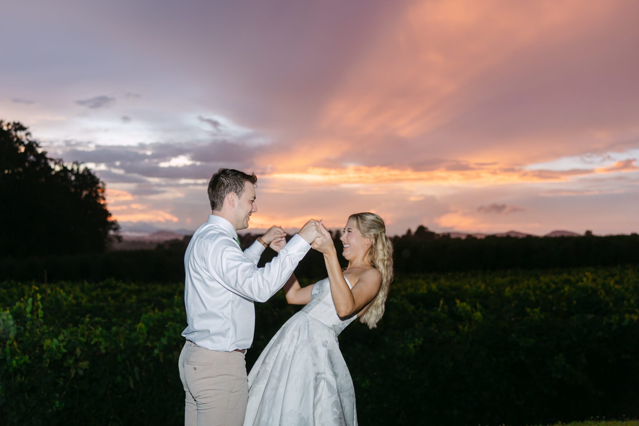 A couple in formal attire holds hands and dances outdoors at sunset, smiling and enjoying the moment against a colorful sky and green landscape.