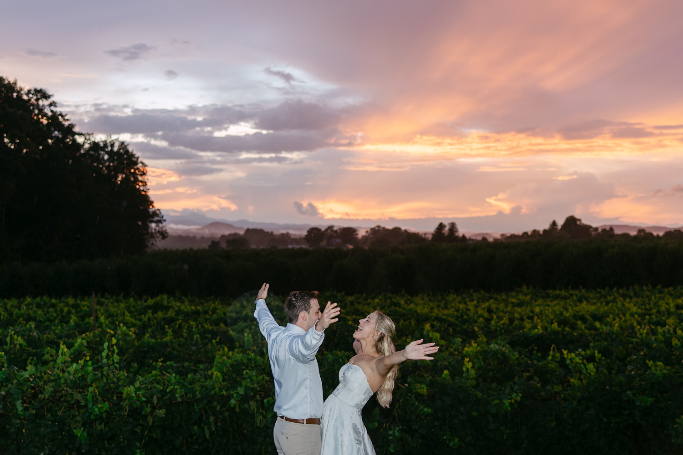 A couple in light clothing stands in the picturesque Marked Tree Vineyards at sunset, joyfully raising their arms and smiling, with dramatic clouds and a colorful sky painting the background.