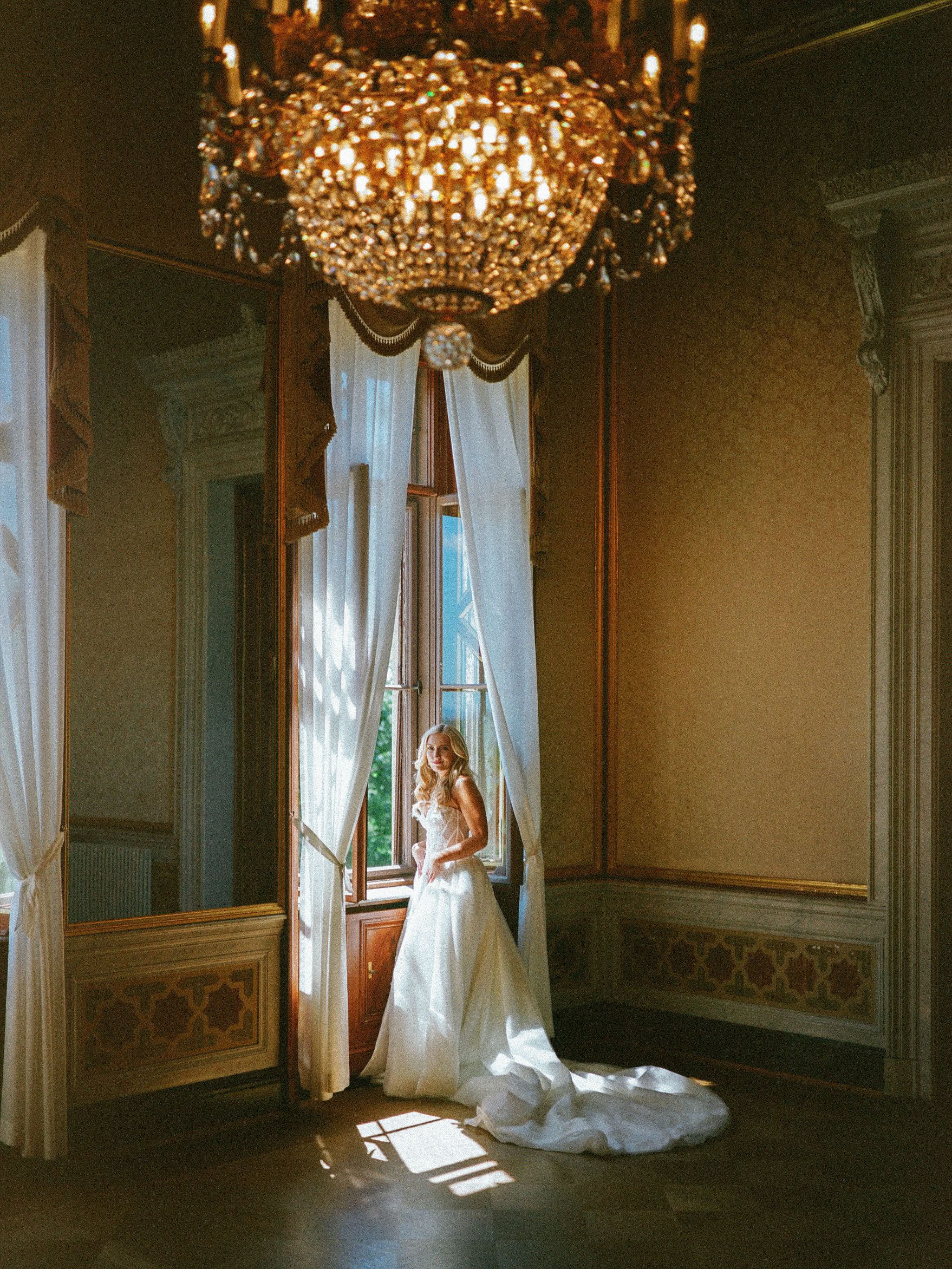 Bride standing at a tall window beneath an ornate chandelier, photographed on film