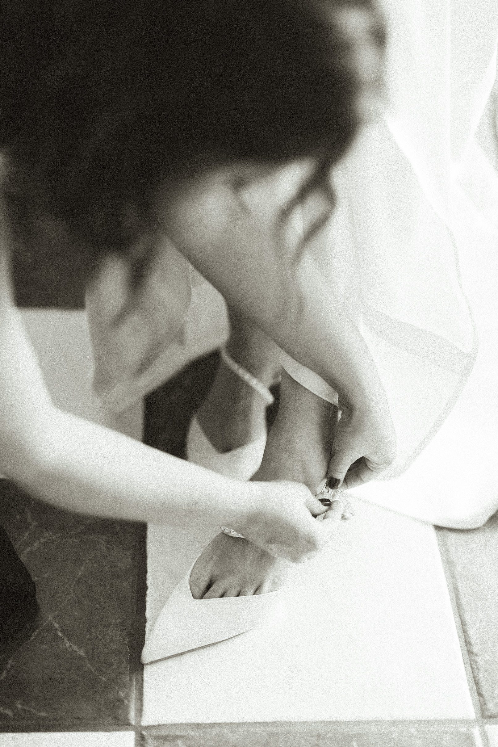 A person in a long white dress helping a bride fasten the strap on her white high-heeled shoe.