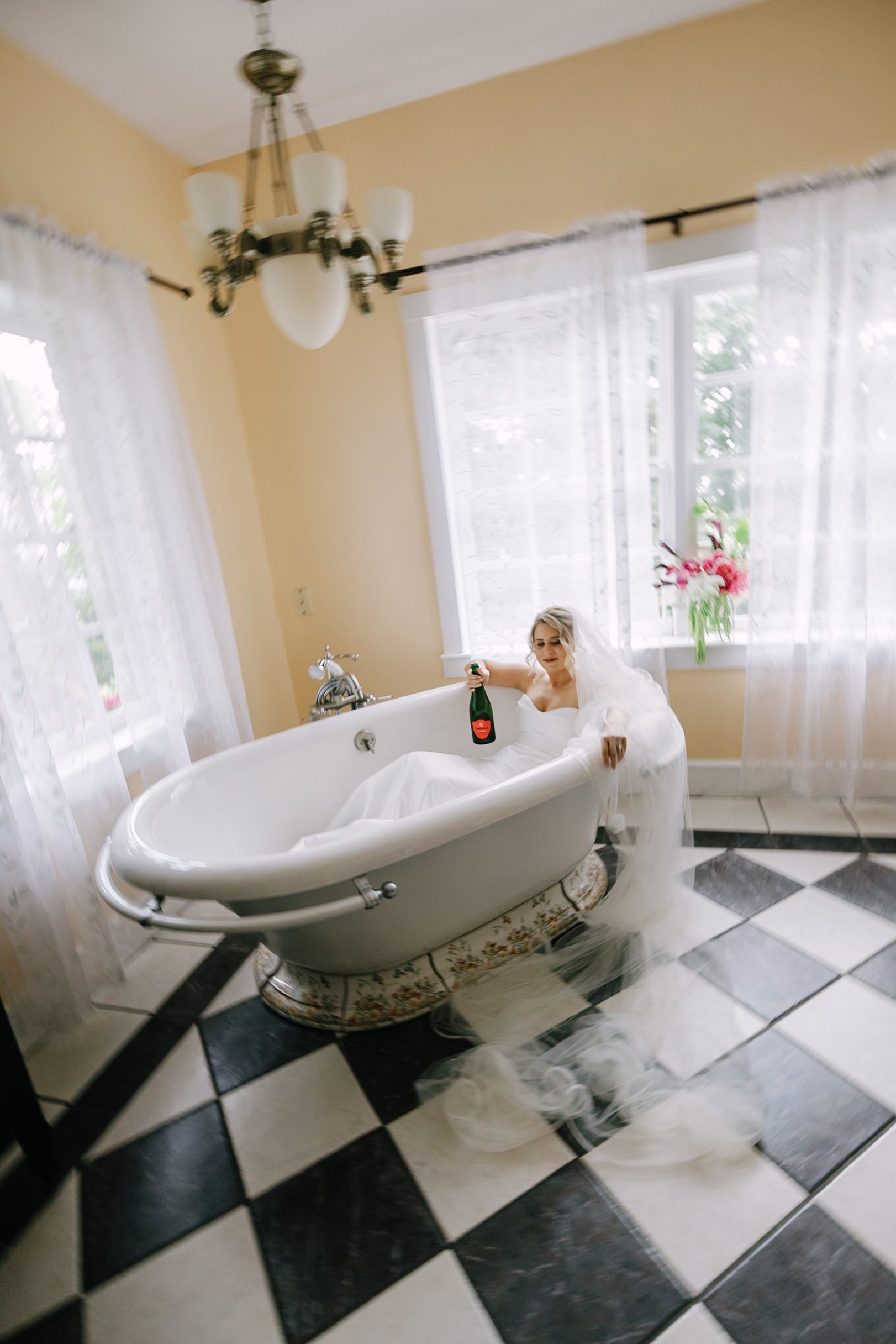A bride in a white wedding dress and veil sits in a bathtub, holding a bottle of champagne.