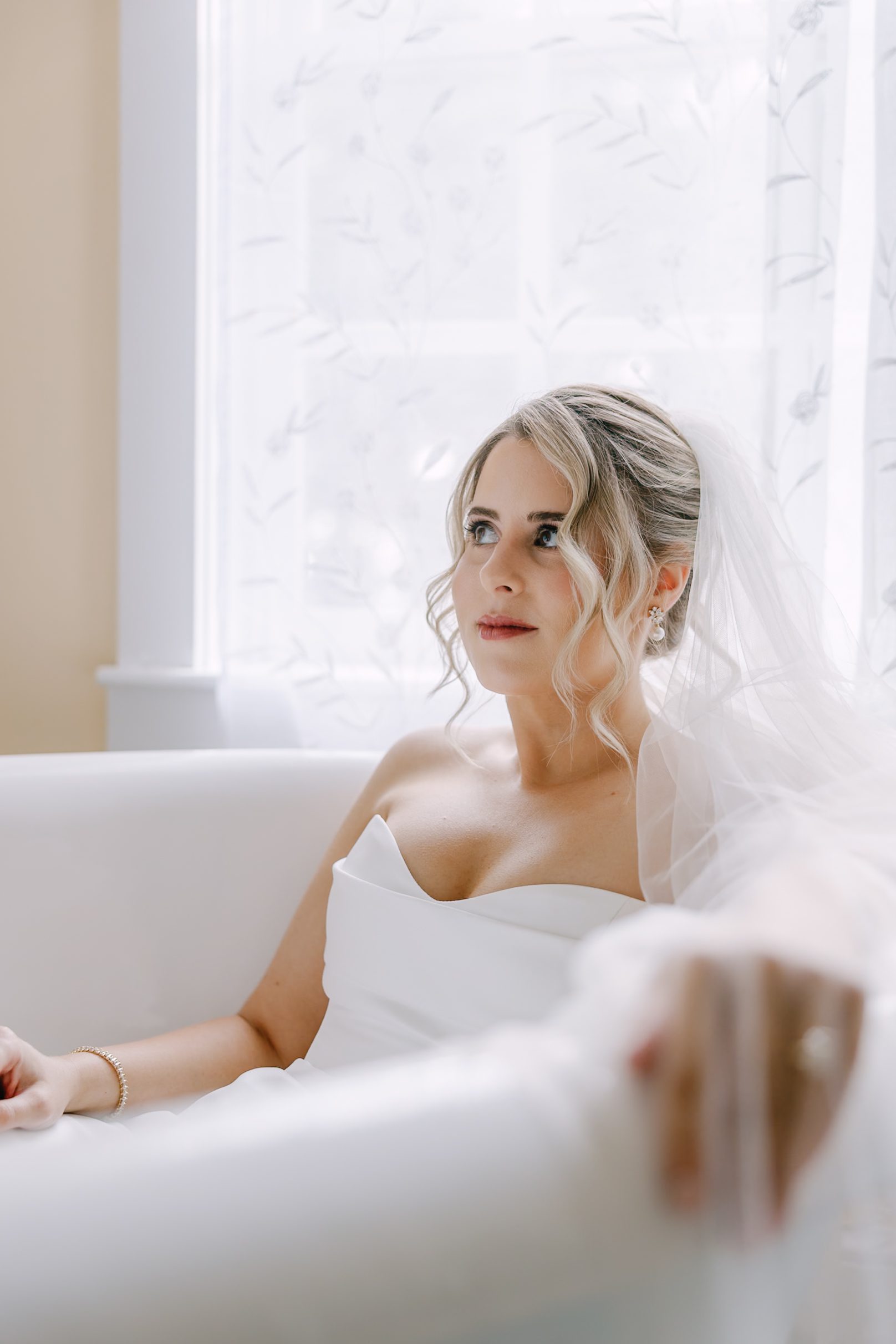 A bride in a strapless white wedding dress and veil sitting in a chair and looking off to the side.