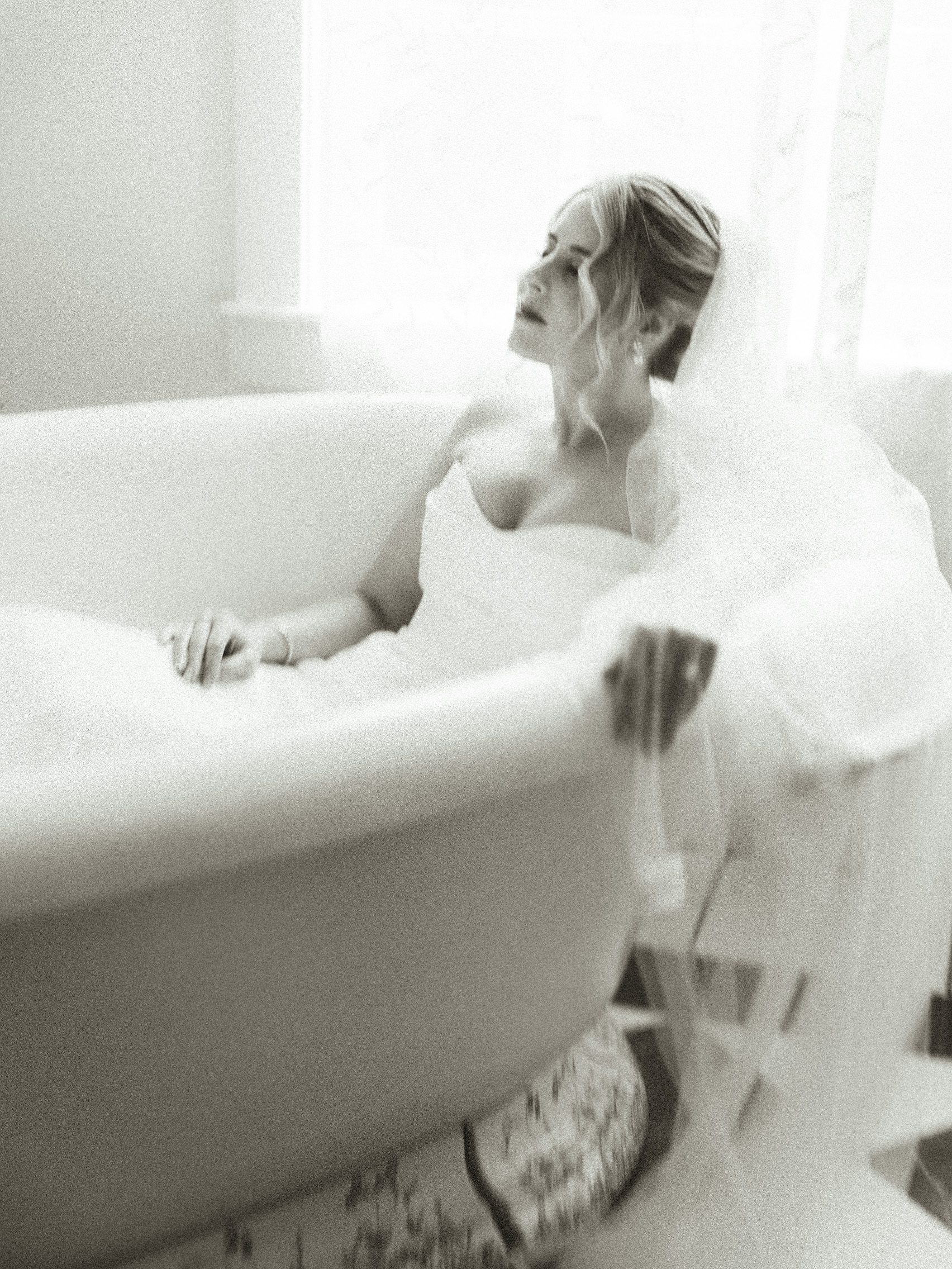 A bride in a white gown and veil sits in a bathtub, looking away with her eyes closed.
