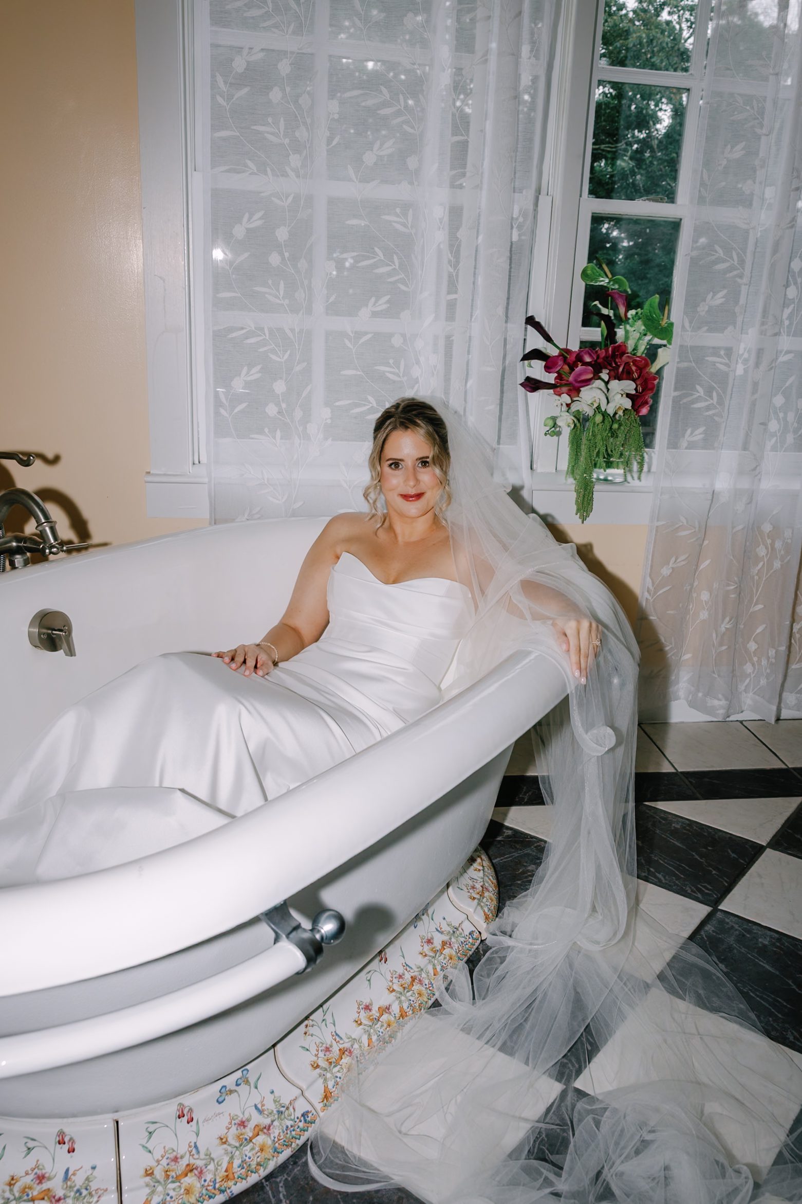 A bride in a strapless white wedding dress and veil sitting in a vintage clawfoot tub.