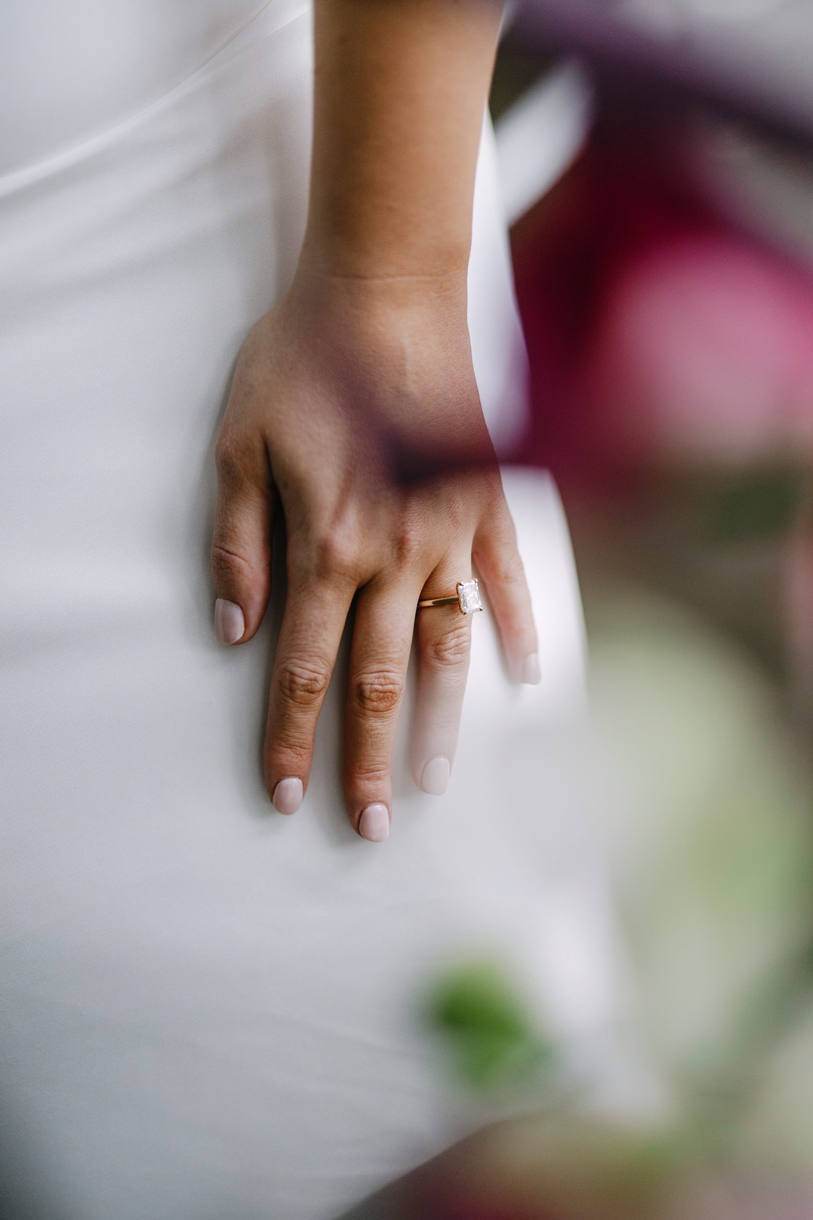 A persons hand with a light-colored manicure resting on a white fabric surface, wearing a gold engagement ring with a rectangular stone.
