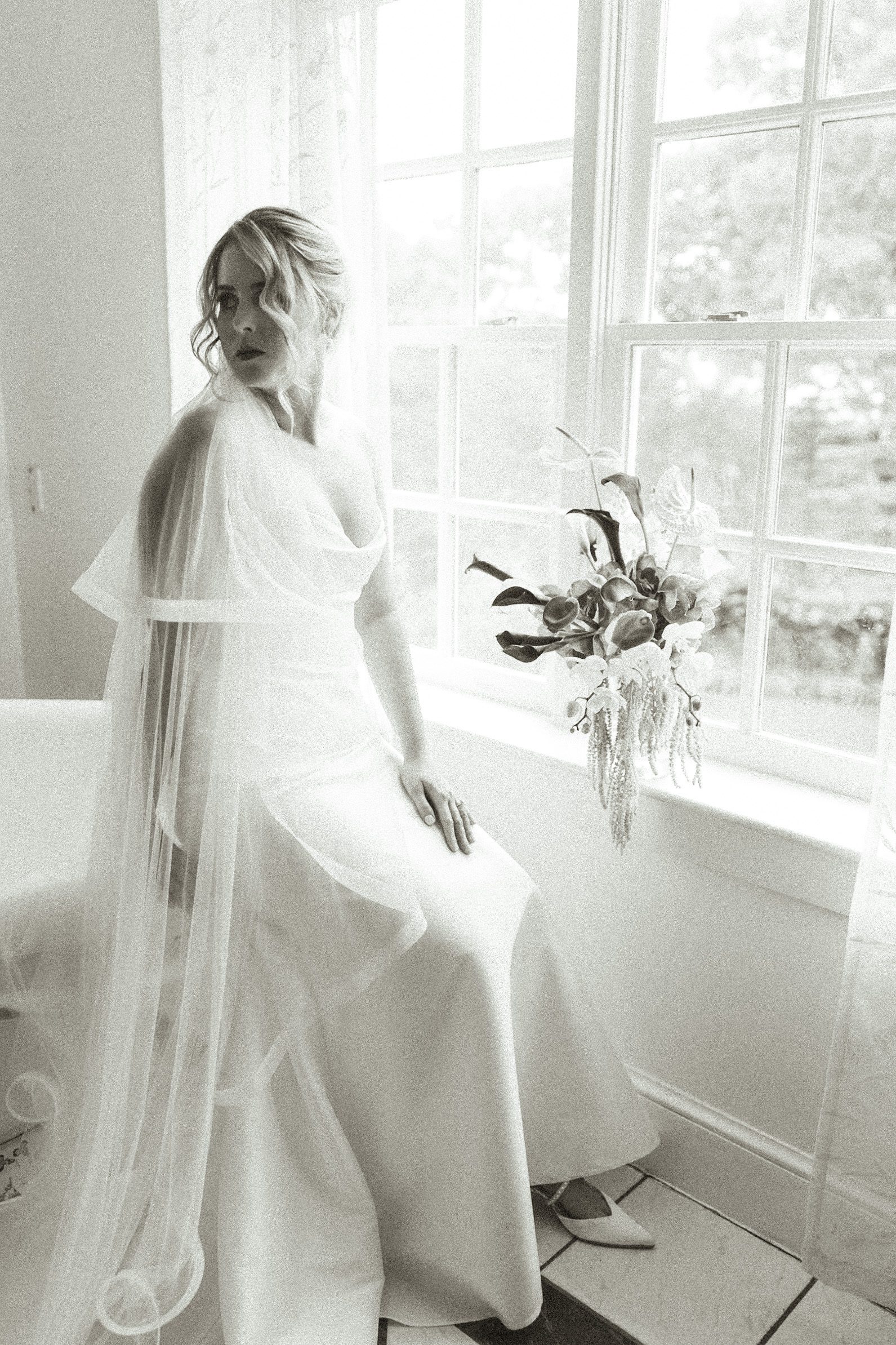 A bride in a gown and veil sits on a window ledge next to a bouquet of flowers.