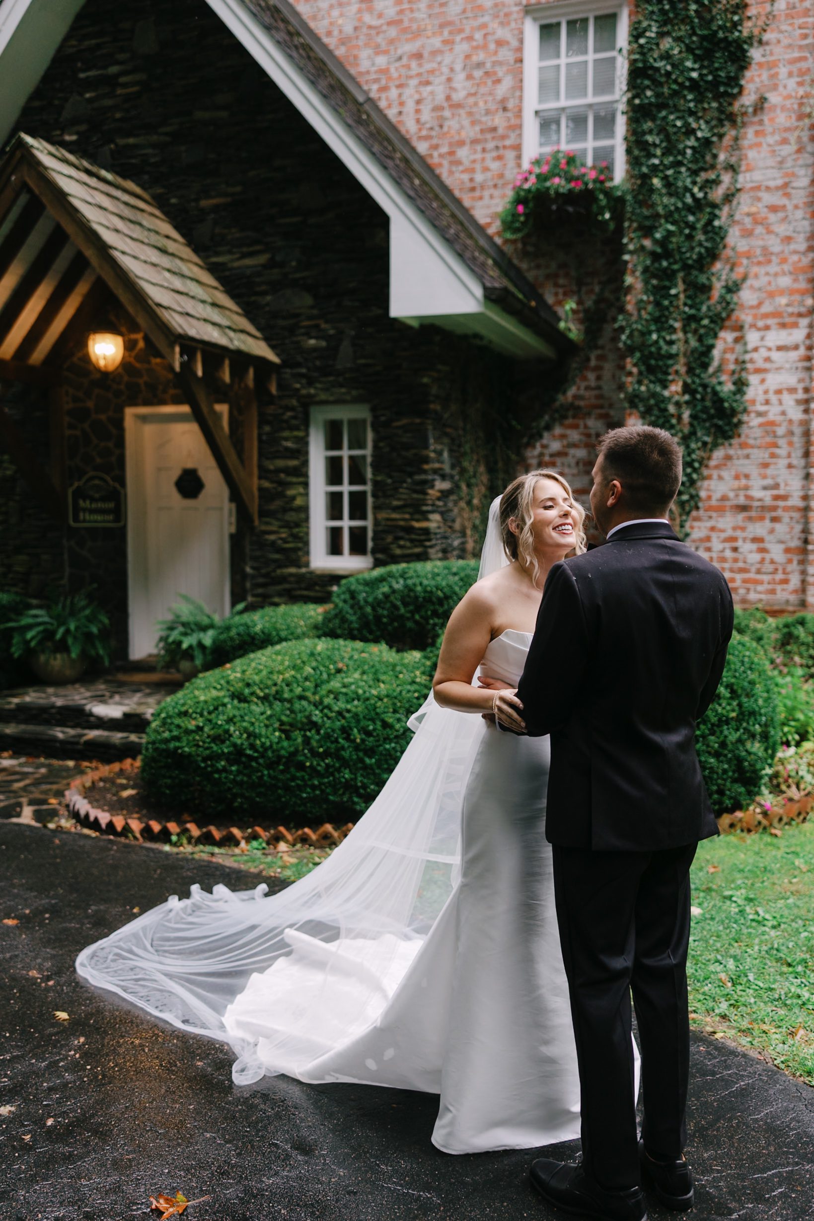 A bride in a white wedding gown and veil smiling at her groom in a black suit while standing outside a stone building.