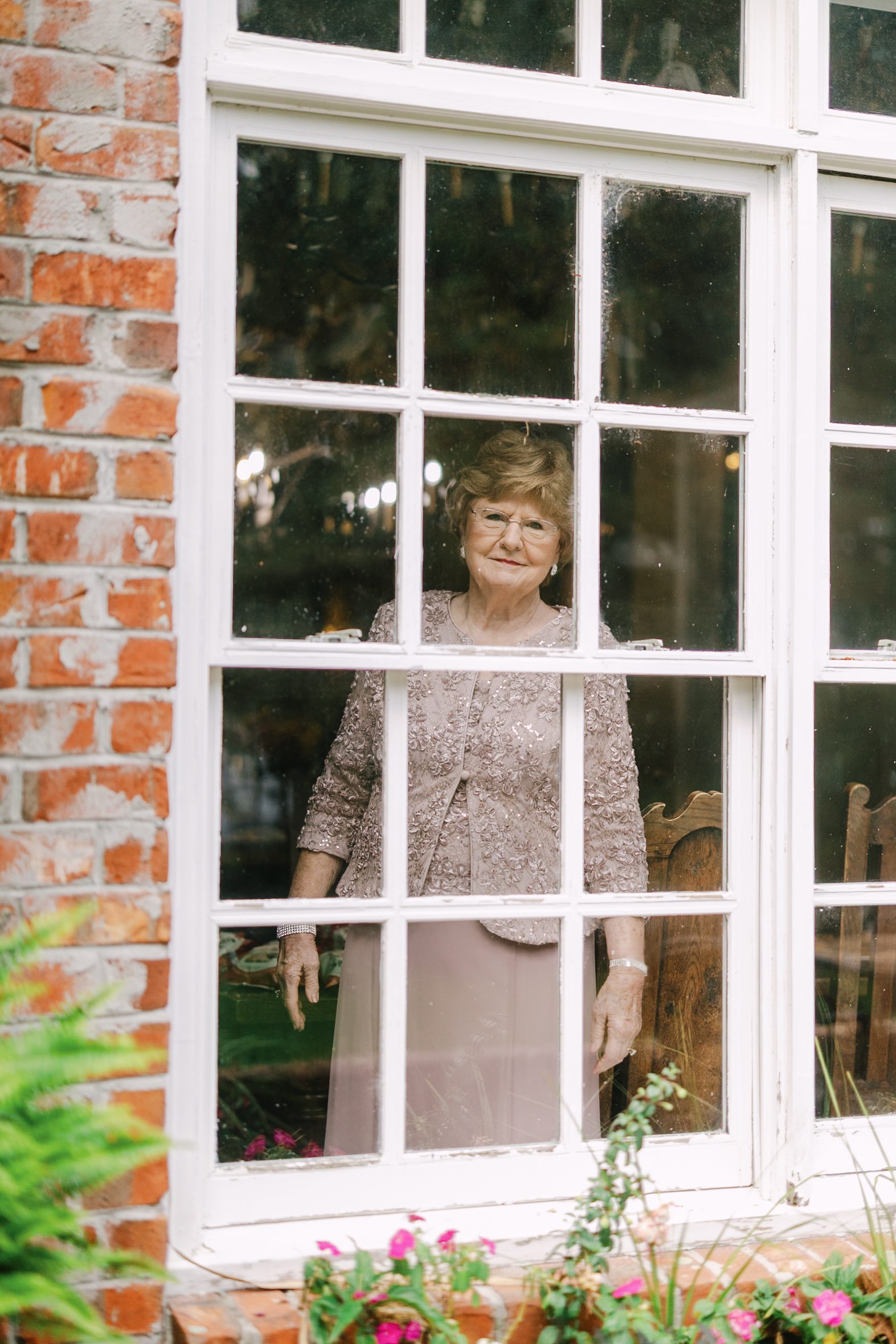 An older woman in a formal matching dress and jacket stands behind a multi-pane window of a brick building.