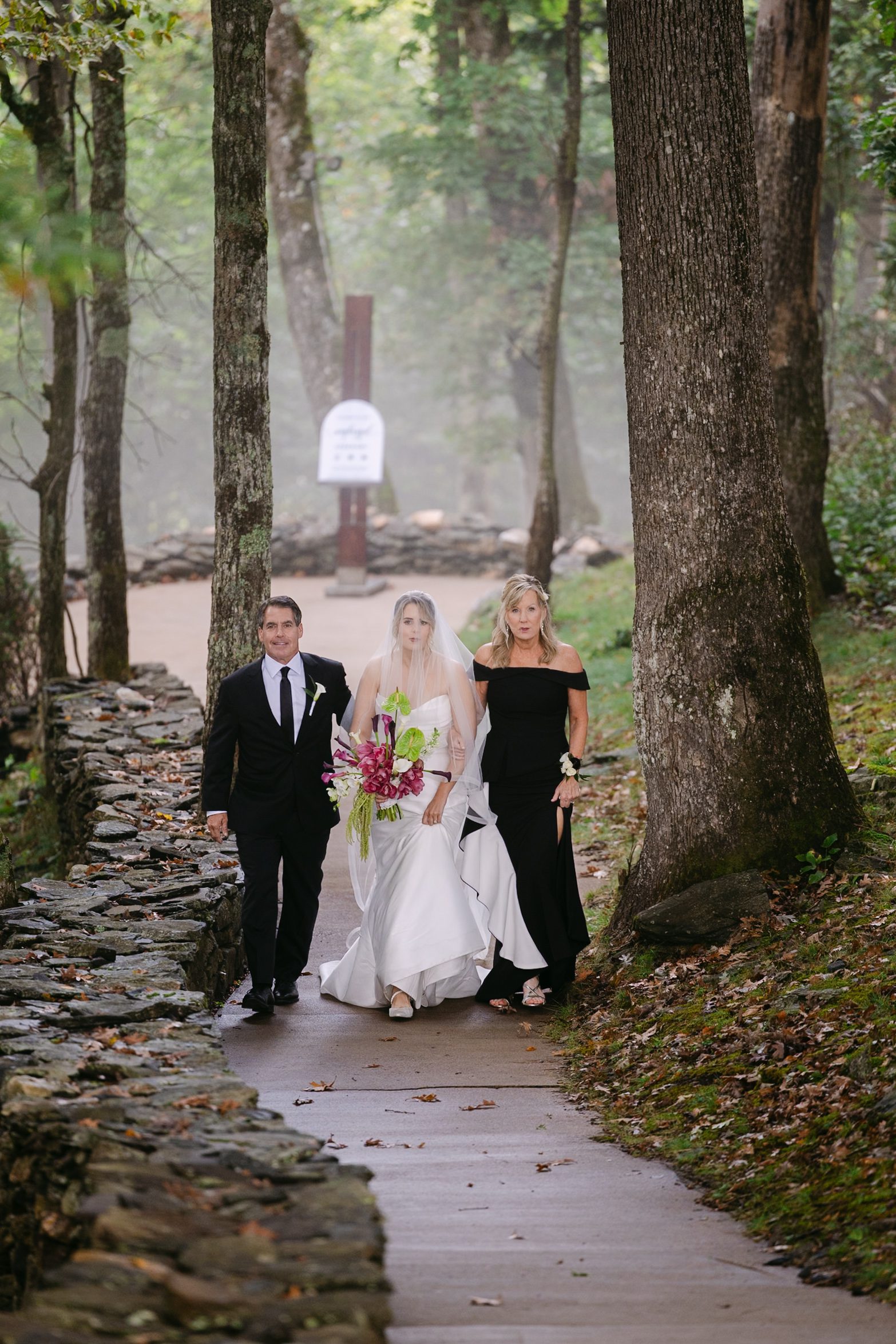 A bride in a white wedding gown walks down a forest path accompanied by her parents.