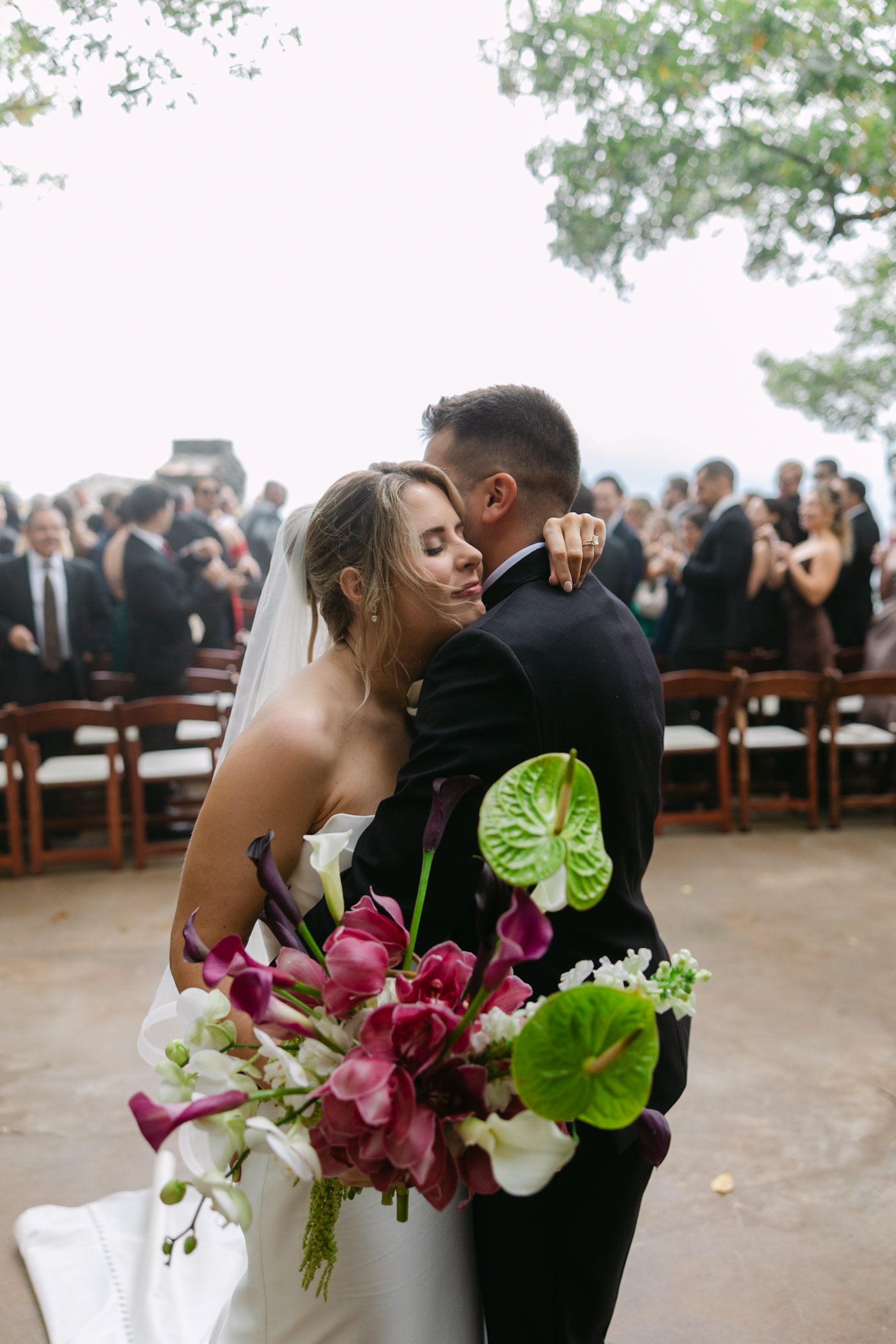 A bride and groom embrace during an outdoor wedding ceremony, with the bride holding a large bouquet of magenta and white flowers.