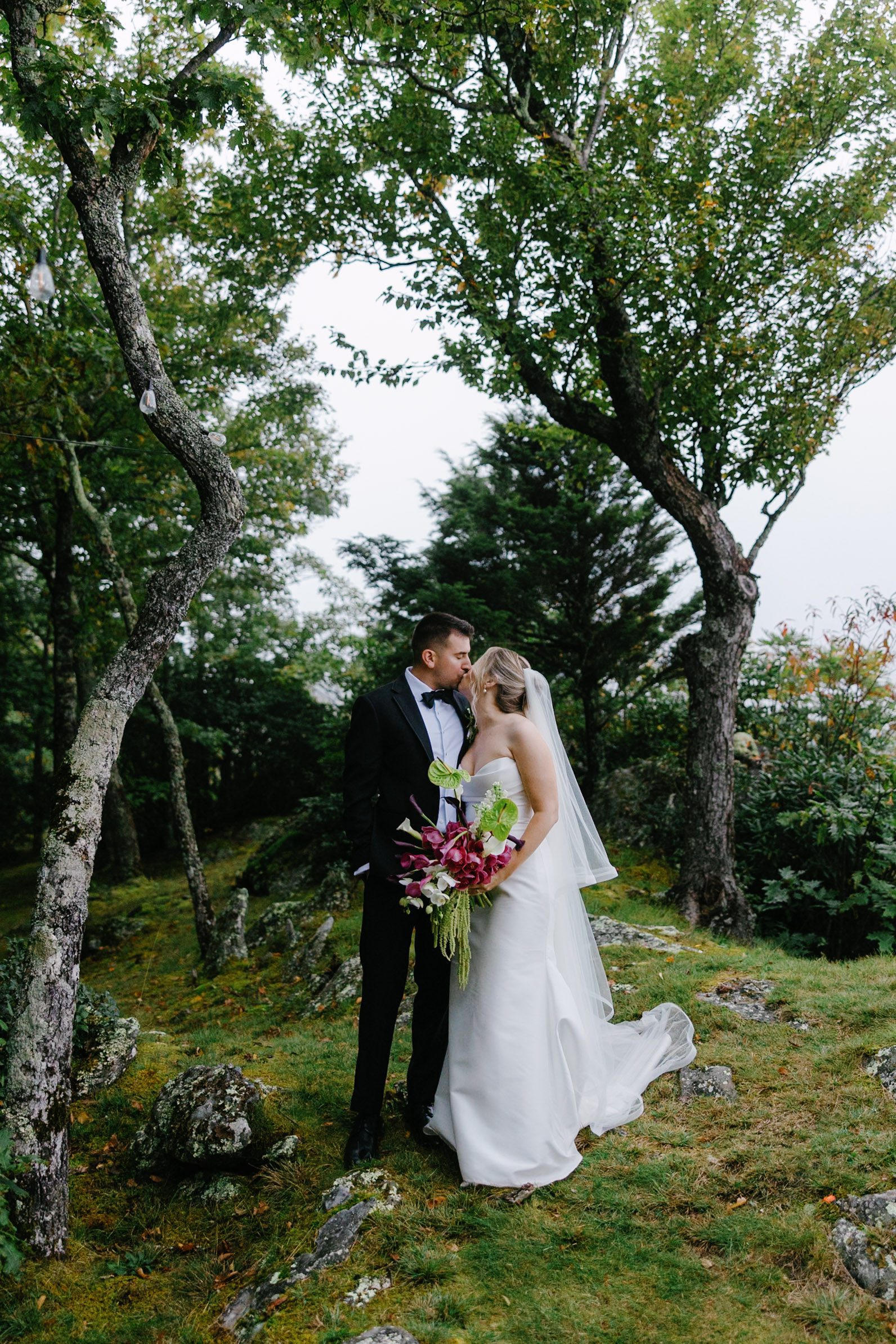 A bride and groom in wedding attire kiss while standing in a wooded area, the bride holding a large bouquet of colorful flowers.