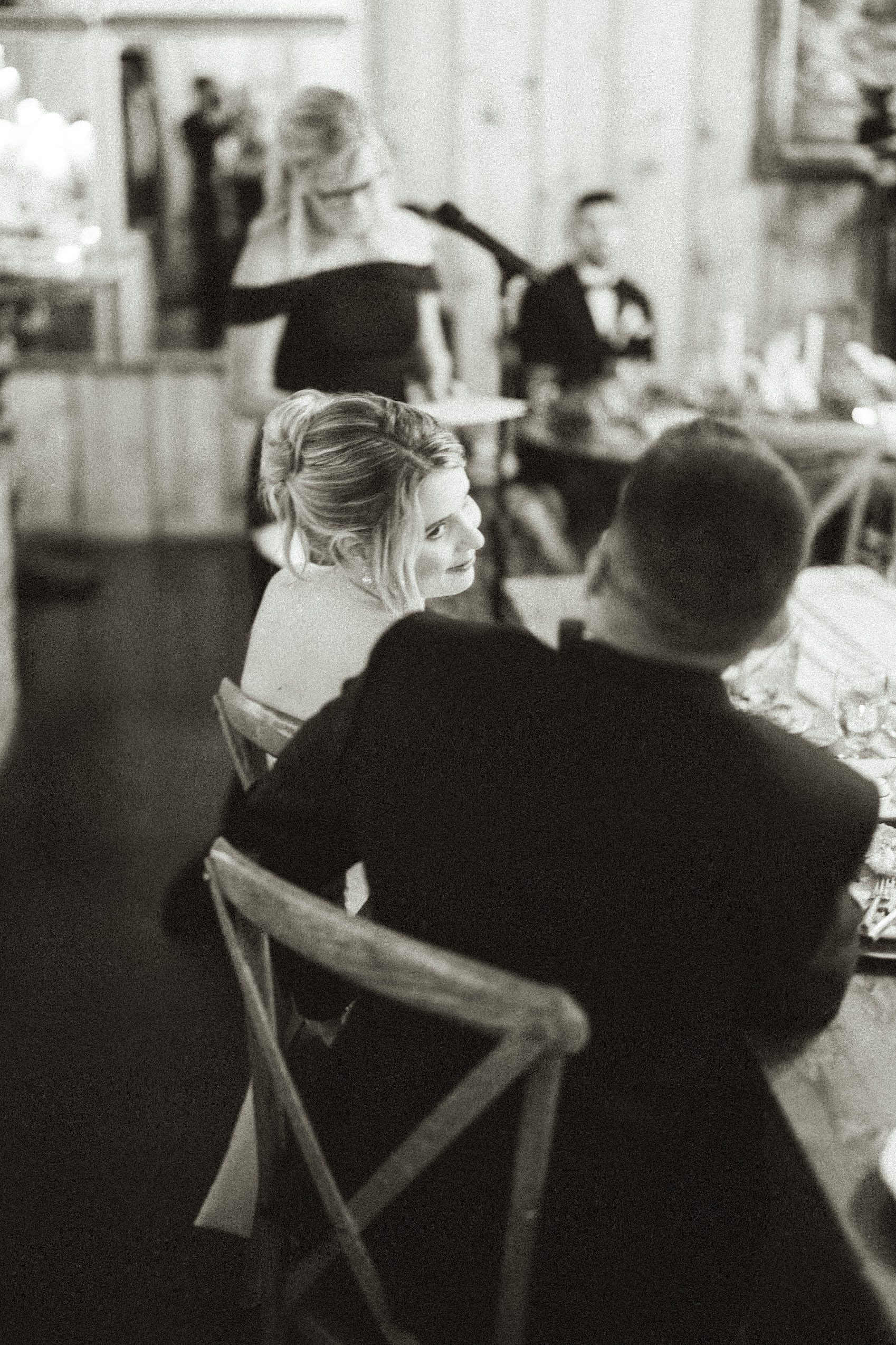 A bride in an updo and a groom in a suit sit at a table looking at each other during a wedding reception.