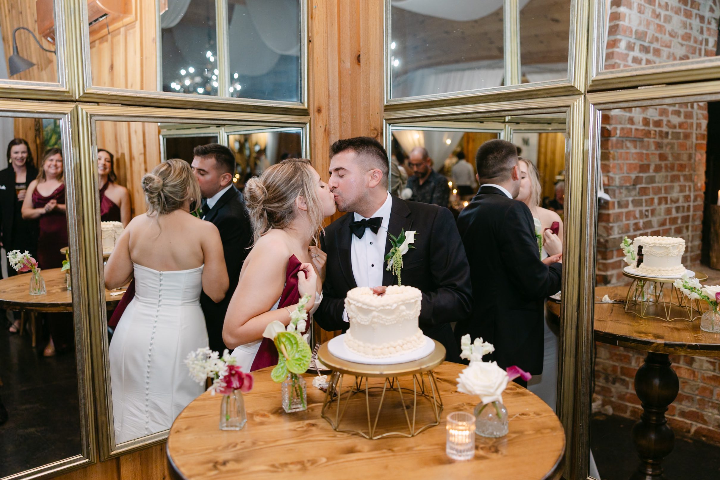 A bride and groom in wedding attire kiss over a small tiered wedding cake on a wooden table, reflected in large, multi-pane mirrors behind them.