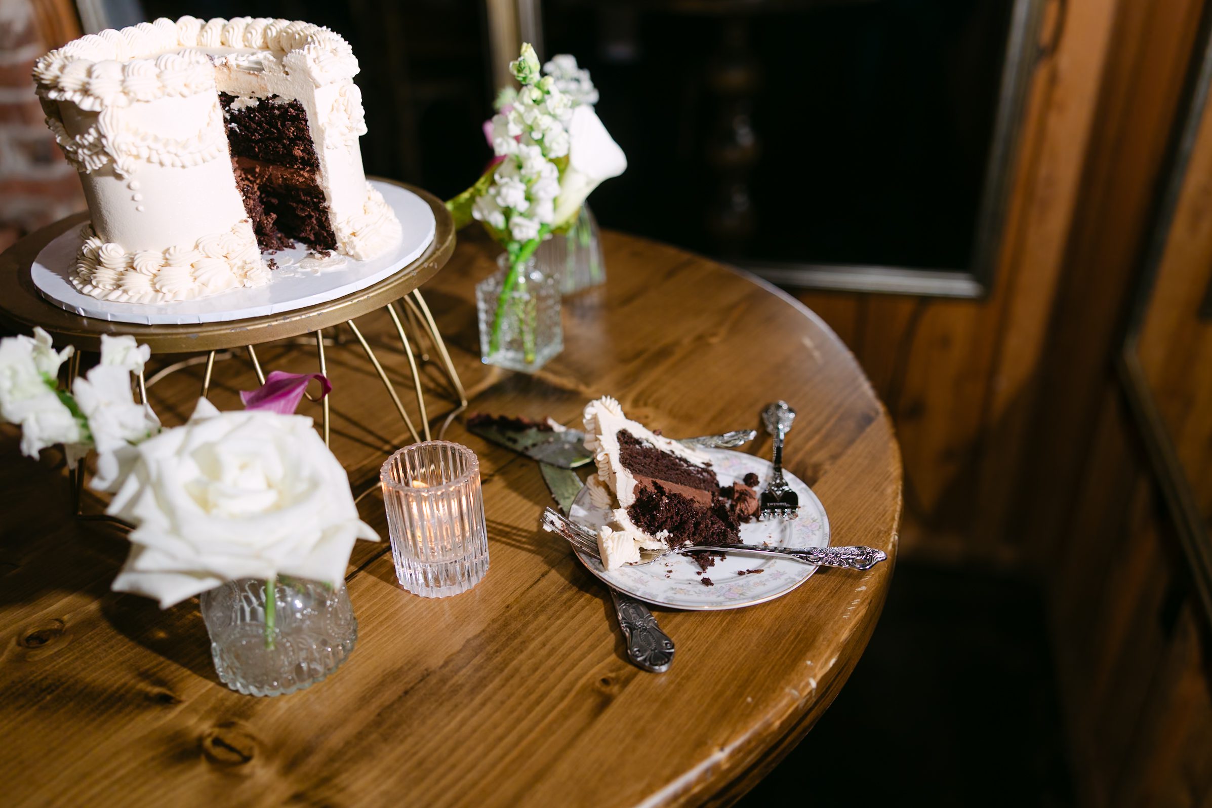 A chocolate cake with white frosting on a tiered stand, with a single slice on a small plate next to a candle and vases of white flowers on a wooden table.
