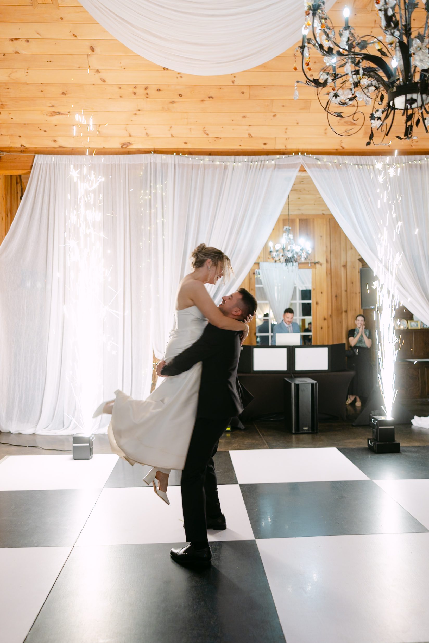 A bride and groom sharing a dance on a black-and-white checkered floor as the groom lifts the bride into his arms, with vertical sparklers glowing in the background.