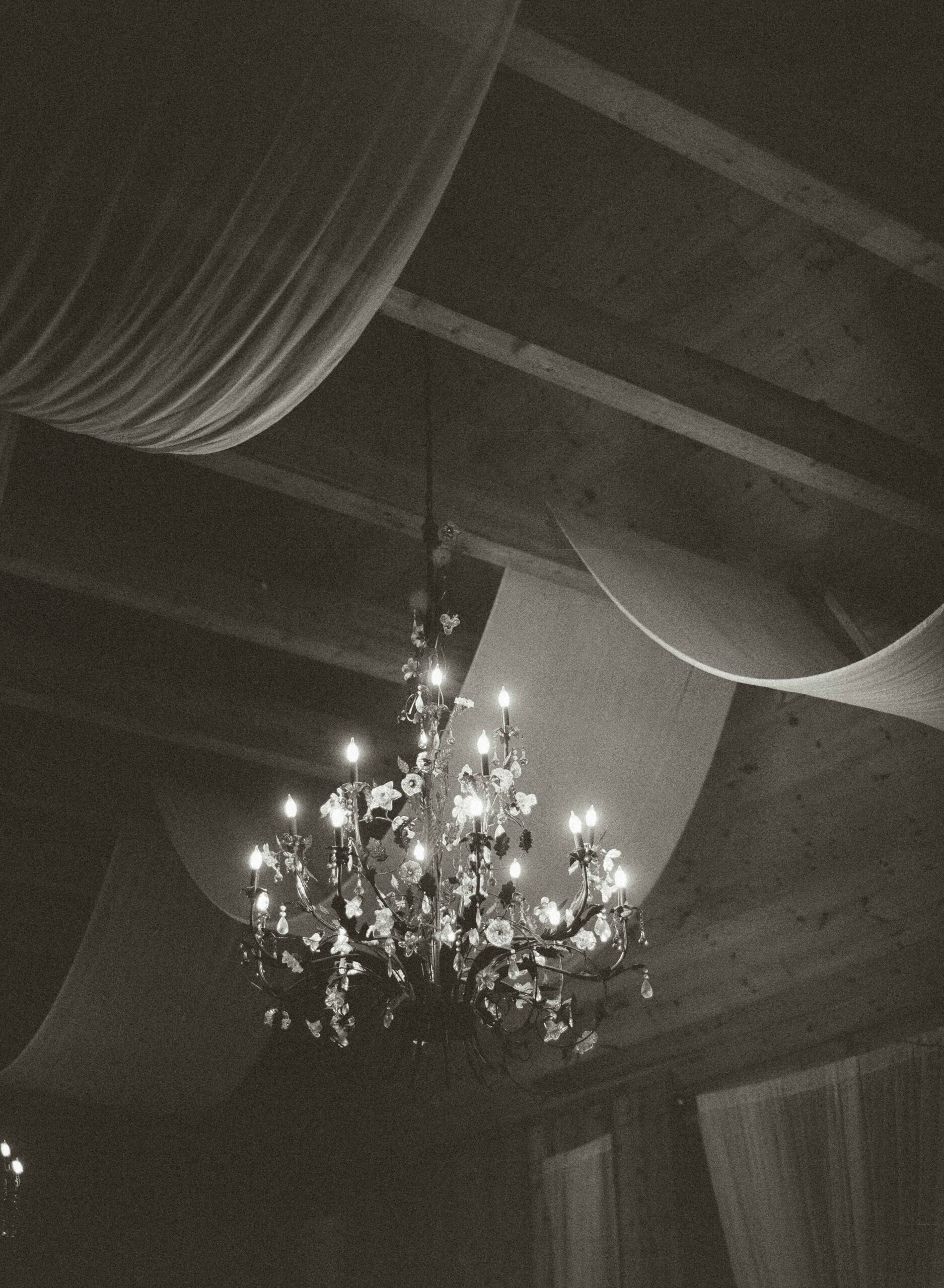A lit chandelier adorned with flowers hangs from a wooden ceiling draped with fabric.