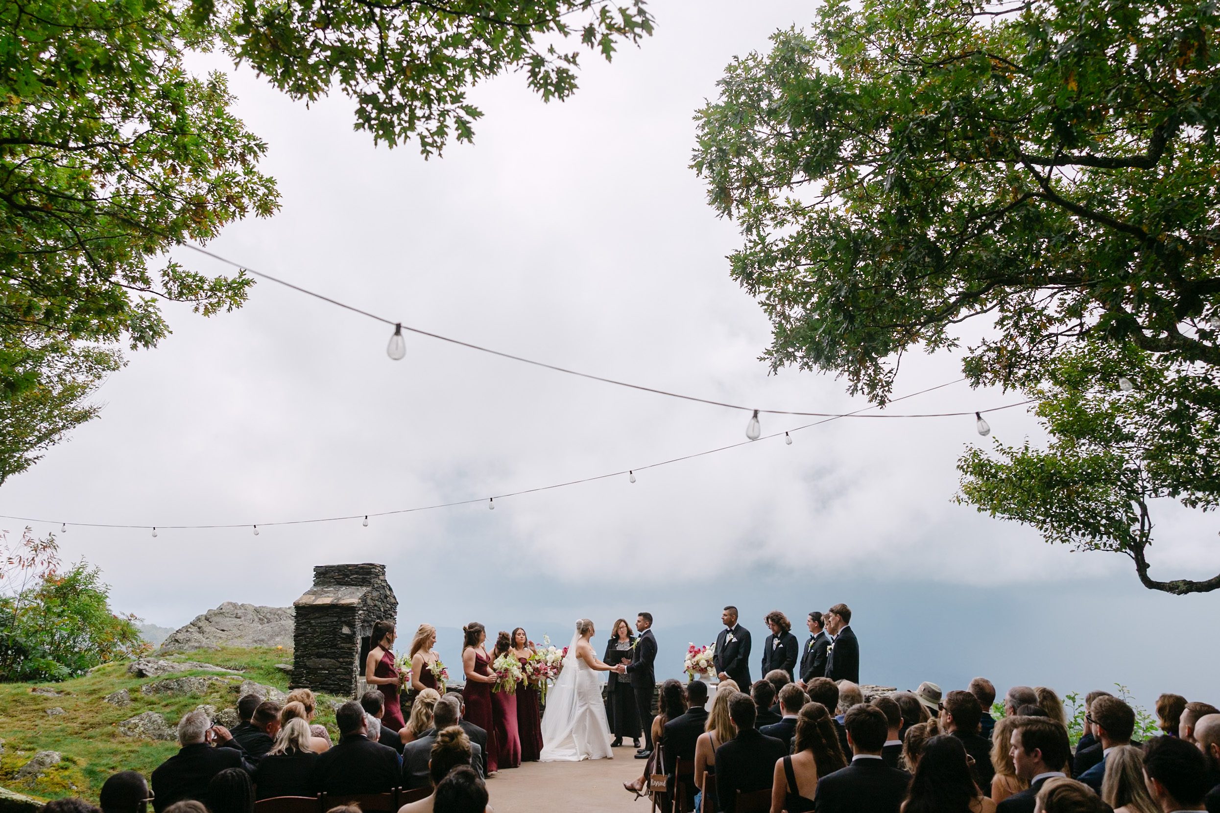 A bride and groom exchange vows at an outdoor cliffside wedding ceremony overlooking a cloudy valley, captured by a Twickenham House film wedding photographer as guests watch from their chairs.