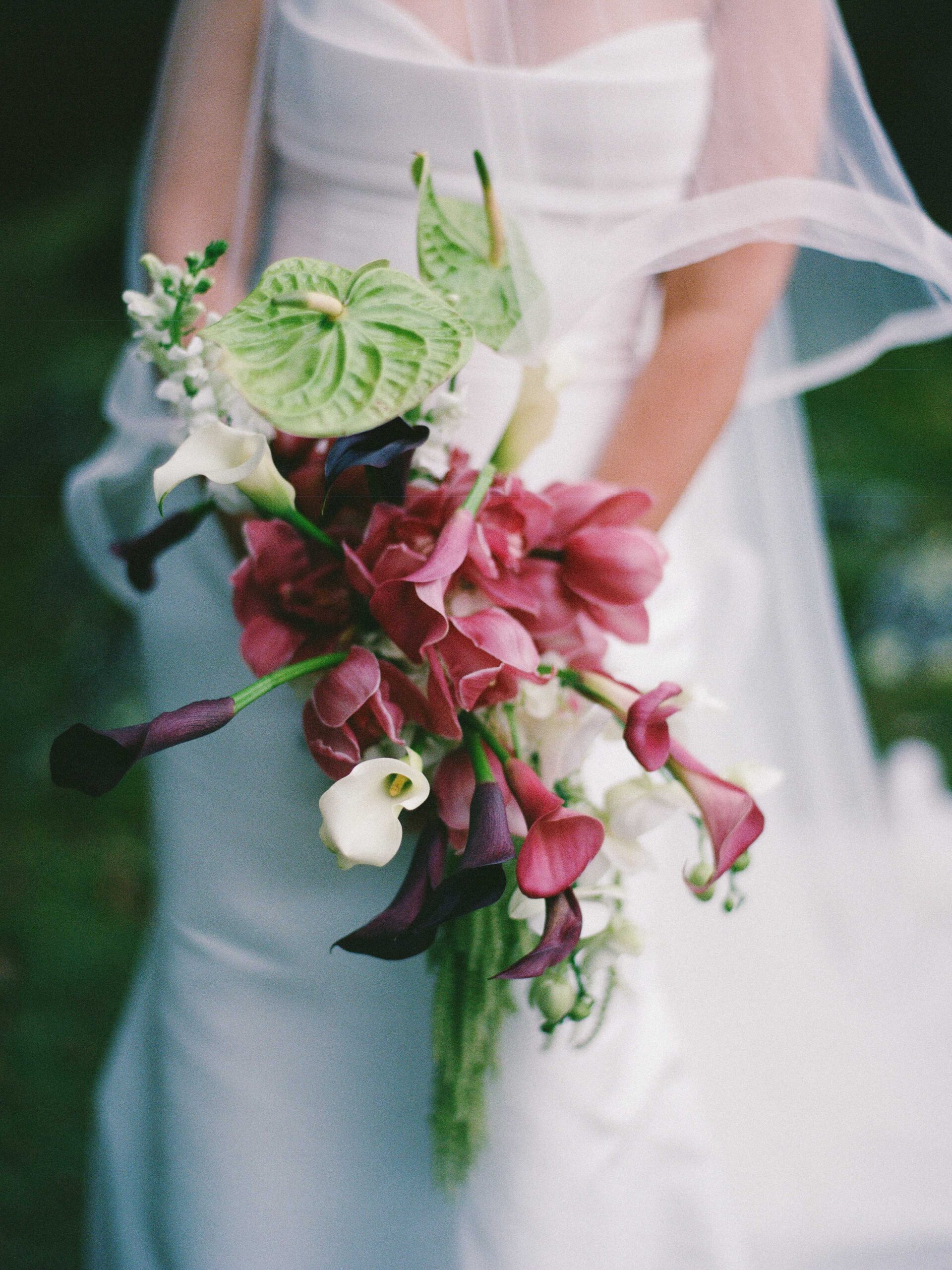 A bride in a white gown and veil holds a bouquet of purple and white calla lilies and pink orchids.