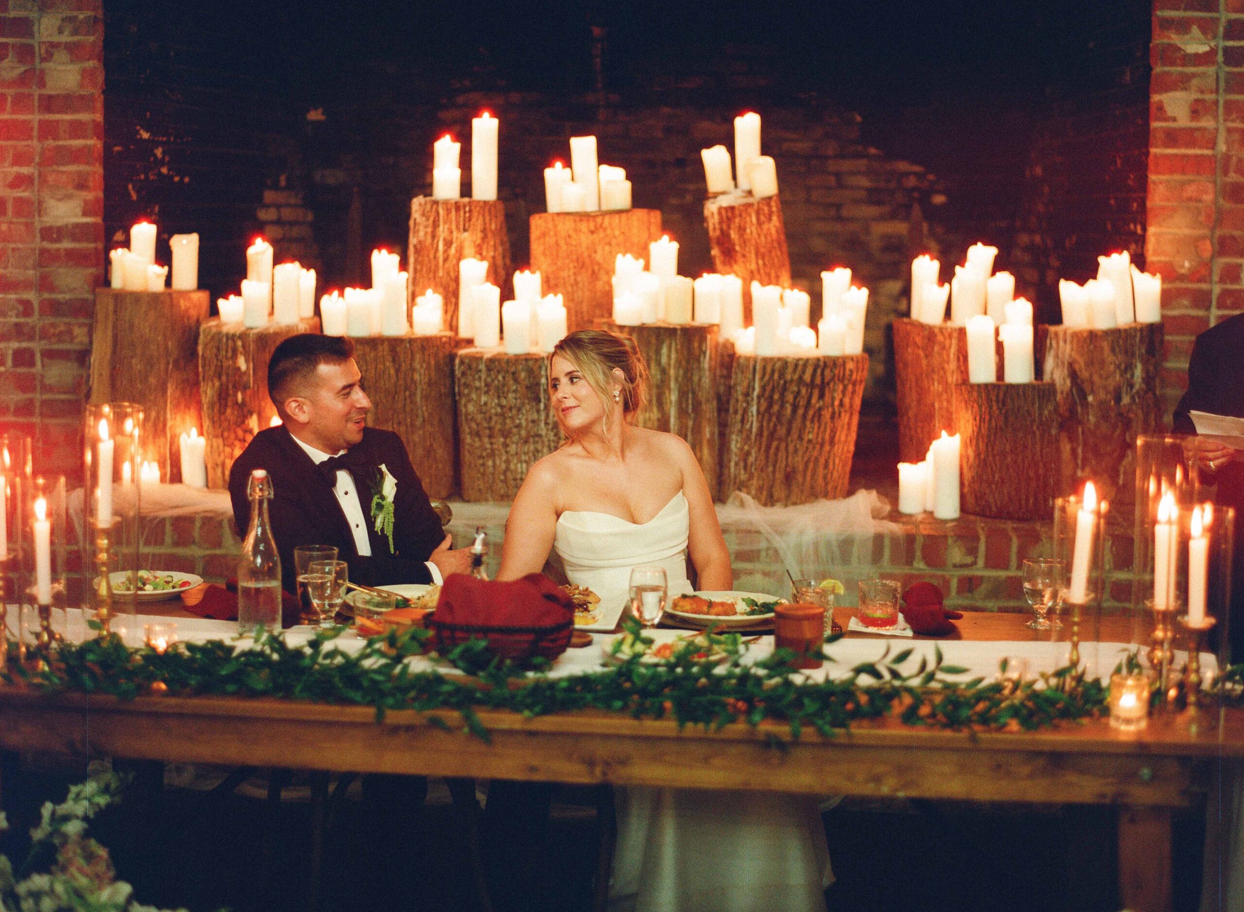 A bride and groom sit at a long table decorated with greenery, smiling at each other in front of a fireplace filled with lit candles on wooden stumps.