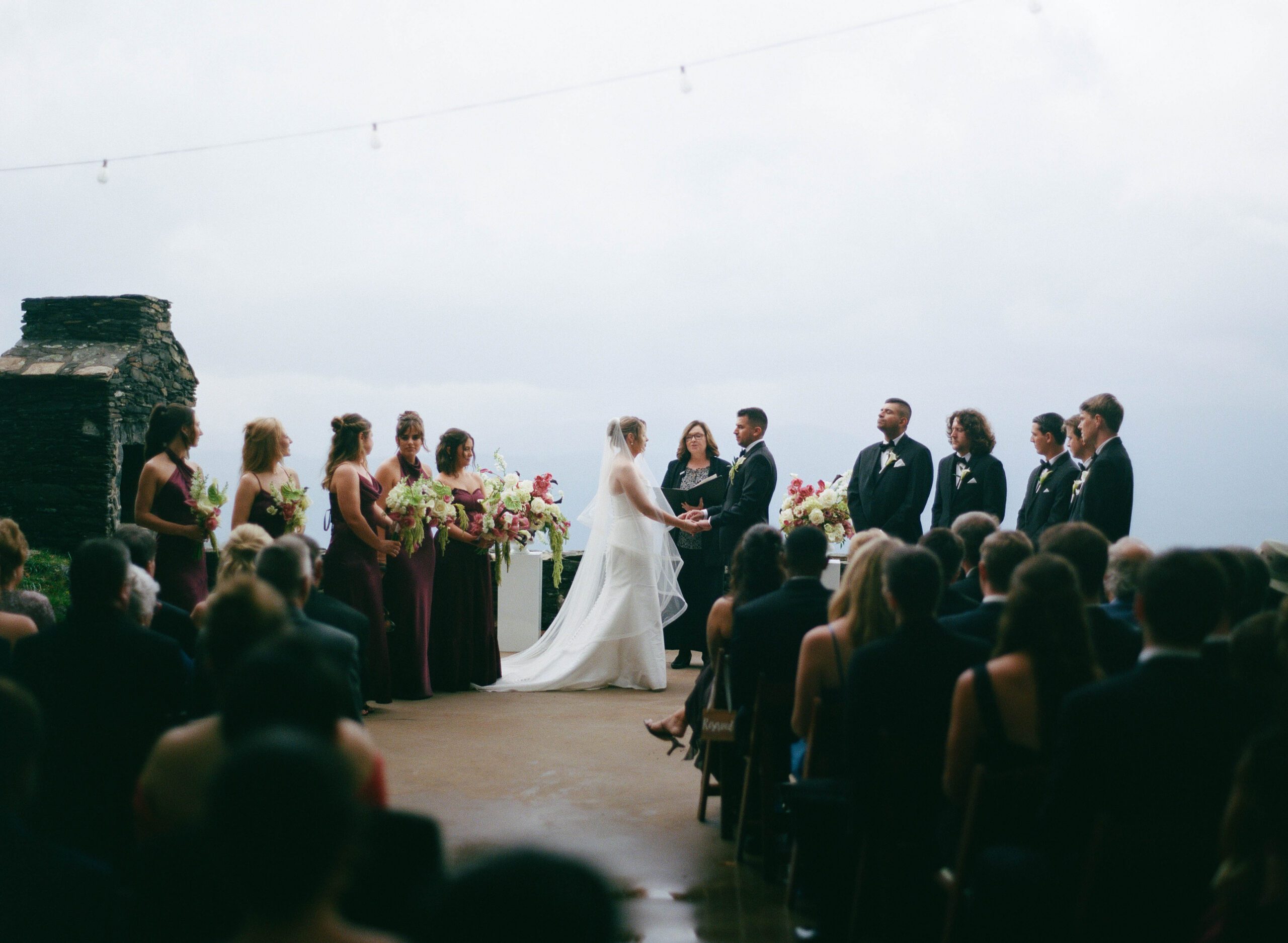 A bride and groom hold hands during an outdoor wedding ceremony, surrounded by their bridesmaids and groomsmen as guests watch from their seats.