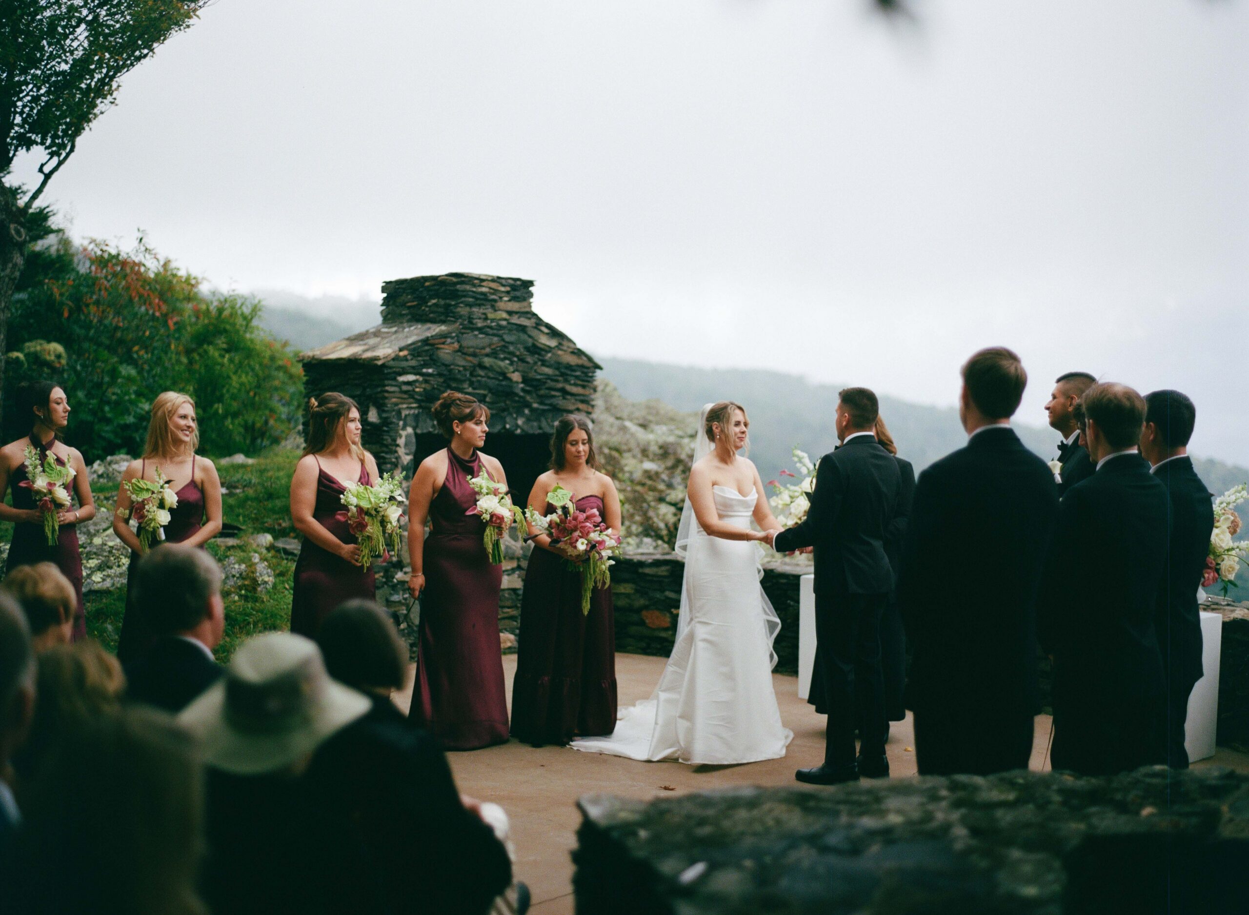A bride and groom hold hands while standing at the altar during an outdoor wedding ceremony, accompanied by bridesmaids in dark red dresses and groomsmen in black suits.