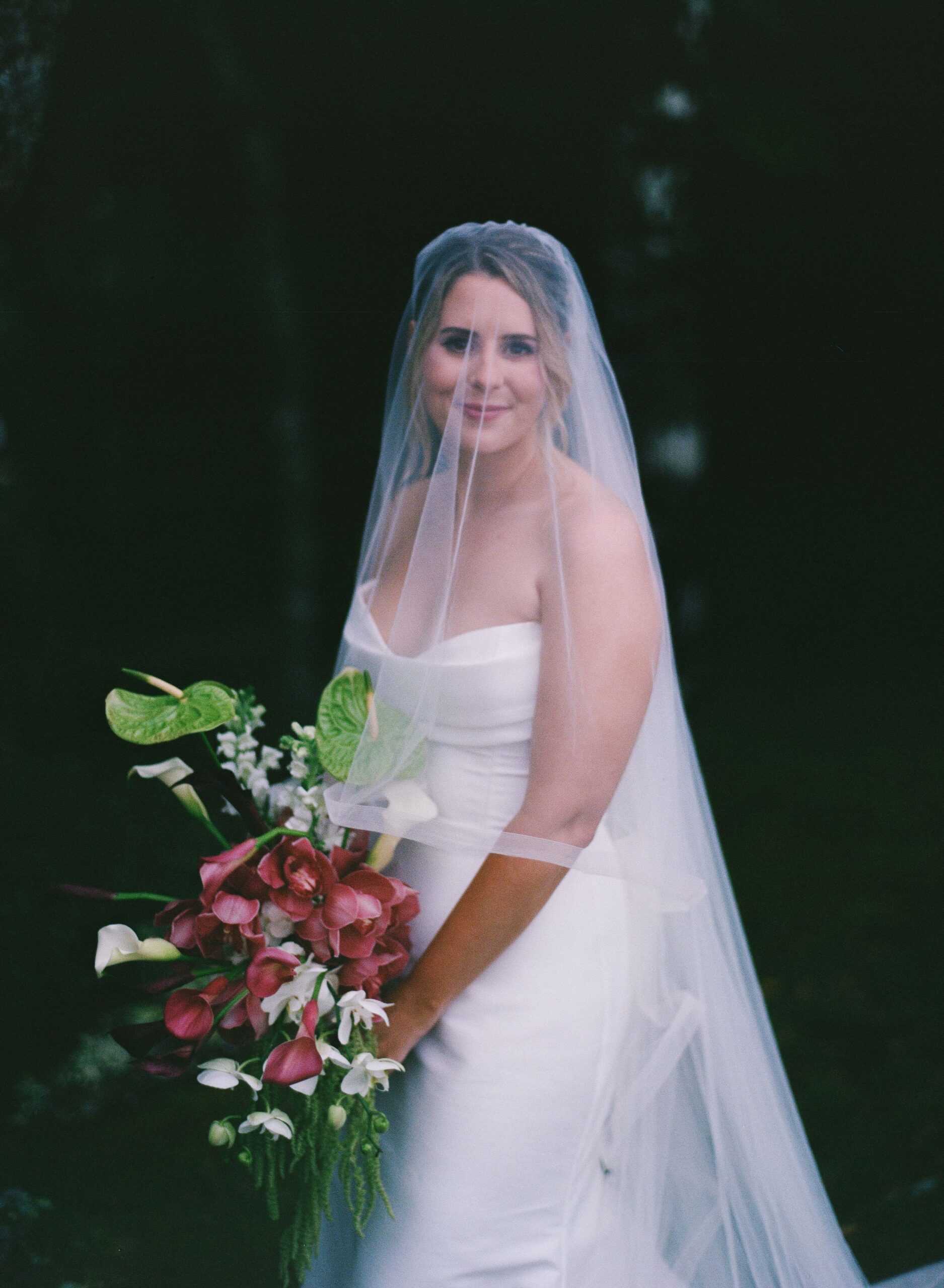 A bride in a strapless white wedding dress and veil holds a large bouquet of red and white flowers.