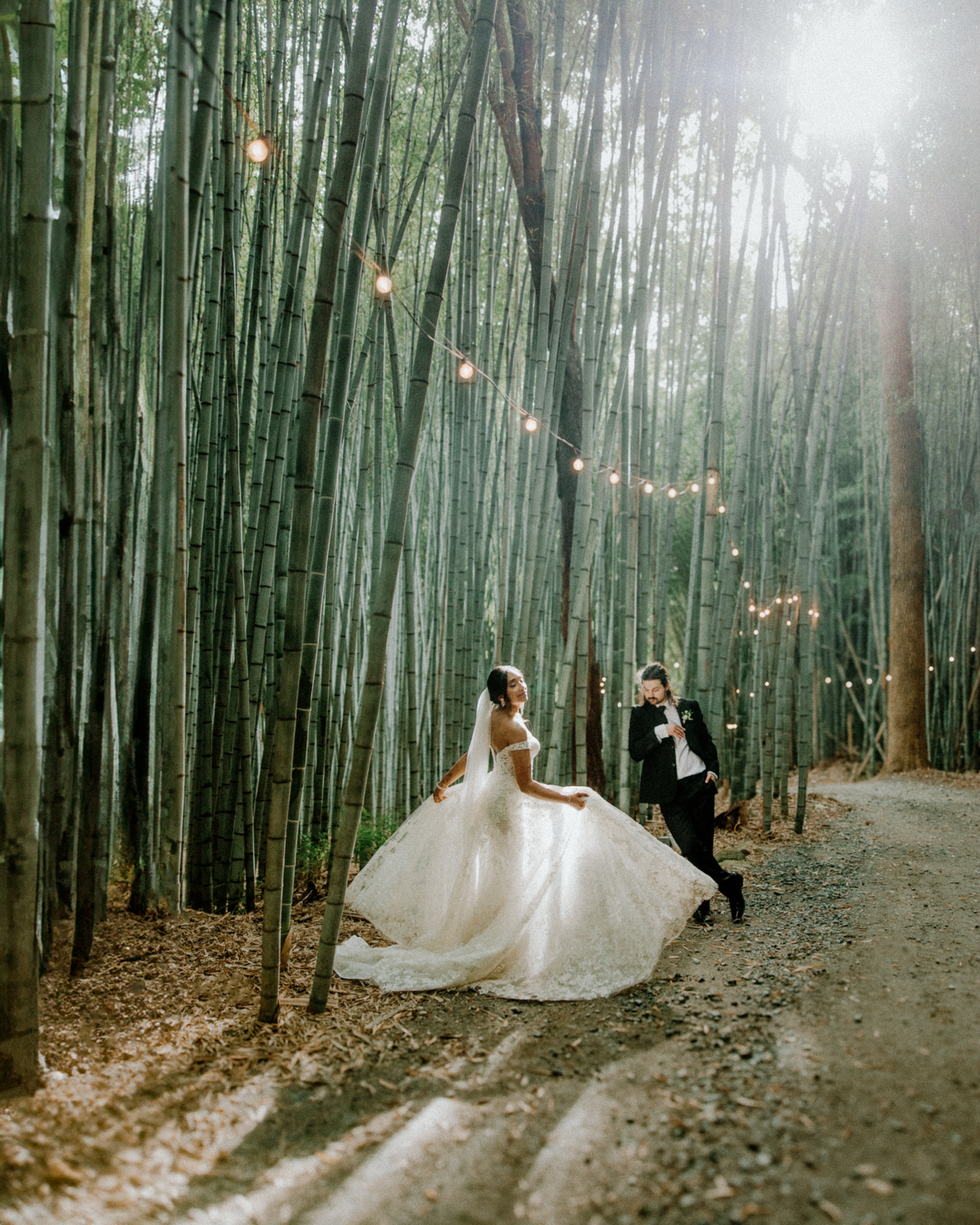 Bride dances in the sunlit bamboo forest of Camelot Meadows wedding venue while groom in tuxedo watches.